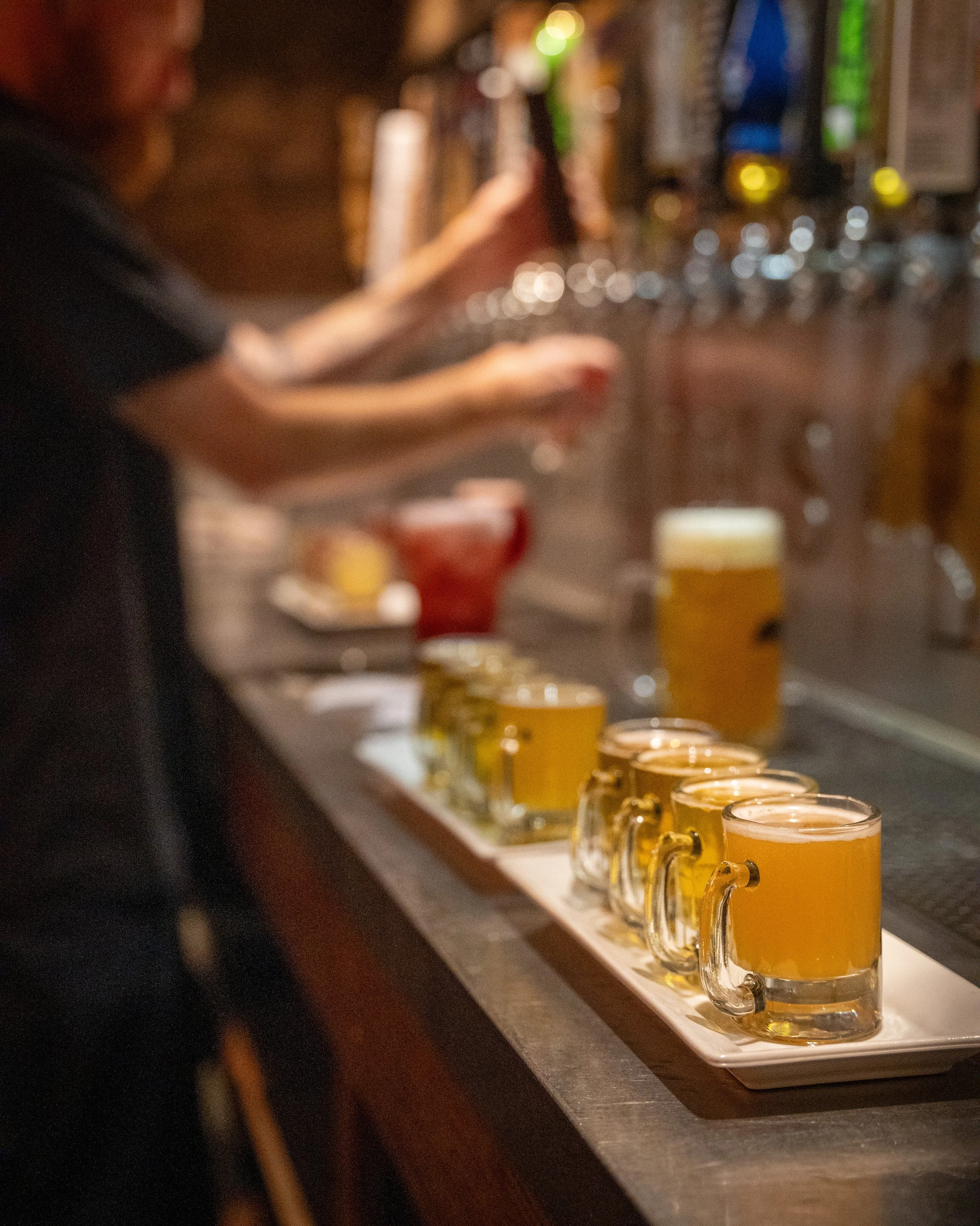 Row of beer mugs filled with beer on a bar counter with a bartender pouring drinks in the background.