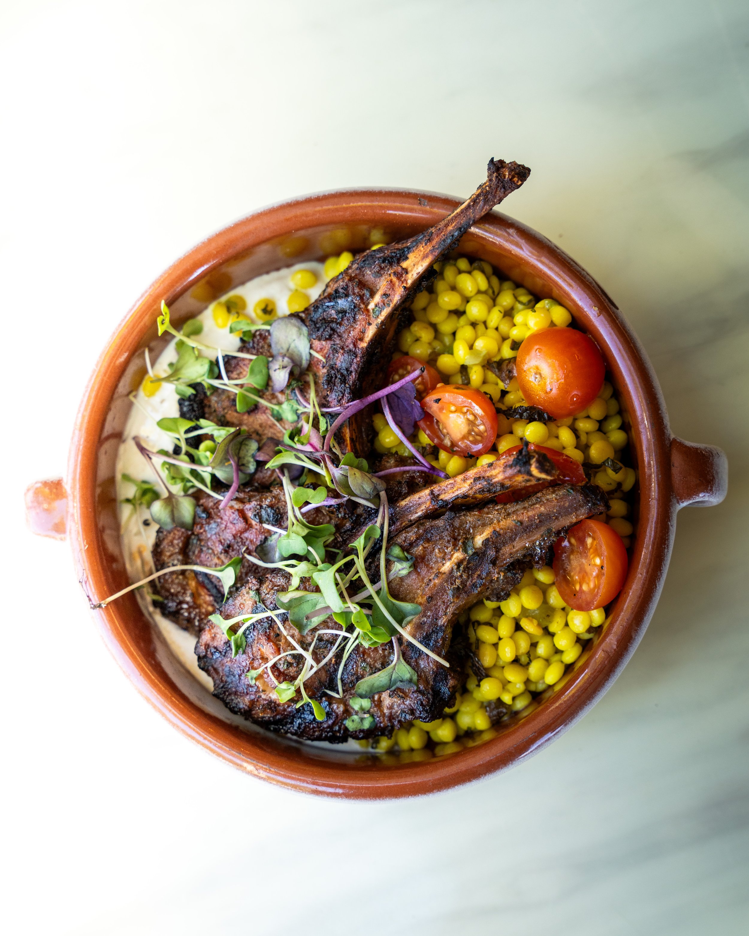 A bowl of grilled lamb chops with microgreens, cherry tomatoes, yellow pearl onions, and creamy sauce.