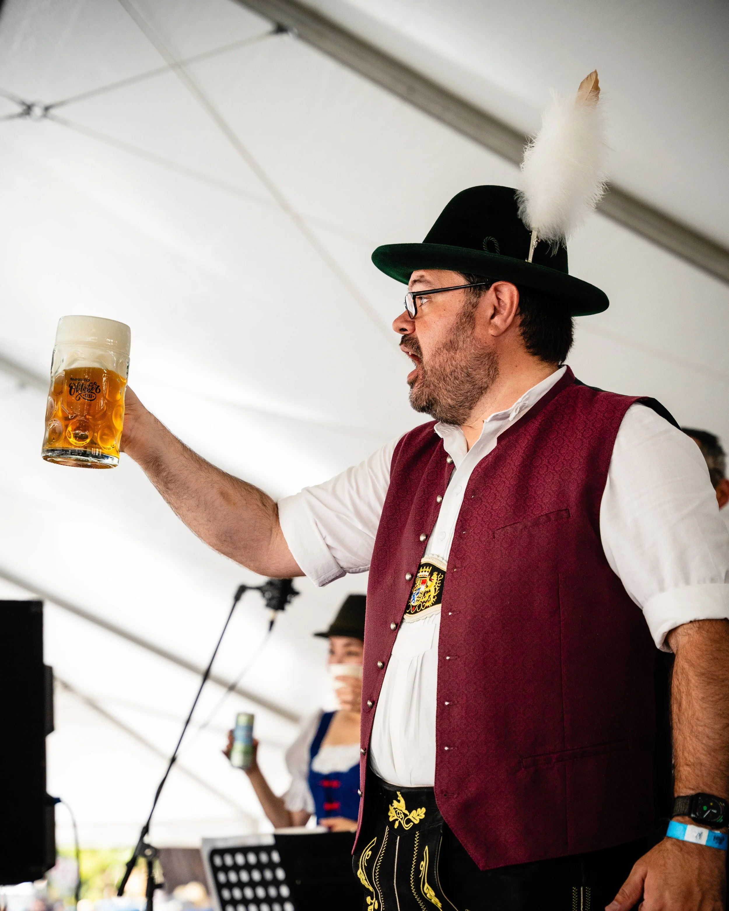 A man dressed in traditional German attire, wearing a black hat with a feather, a white shirt, and a red vest, holding up a large glass of beer during a celebration or festival.