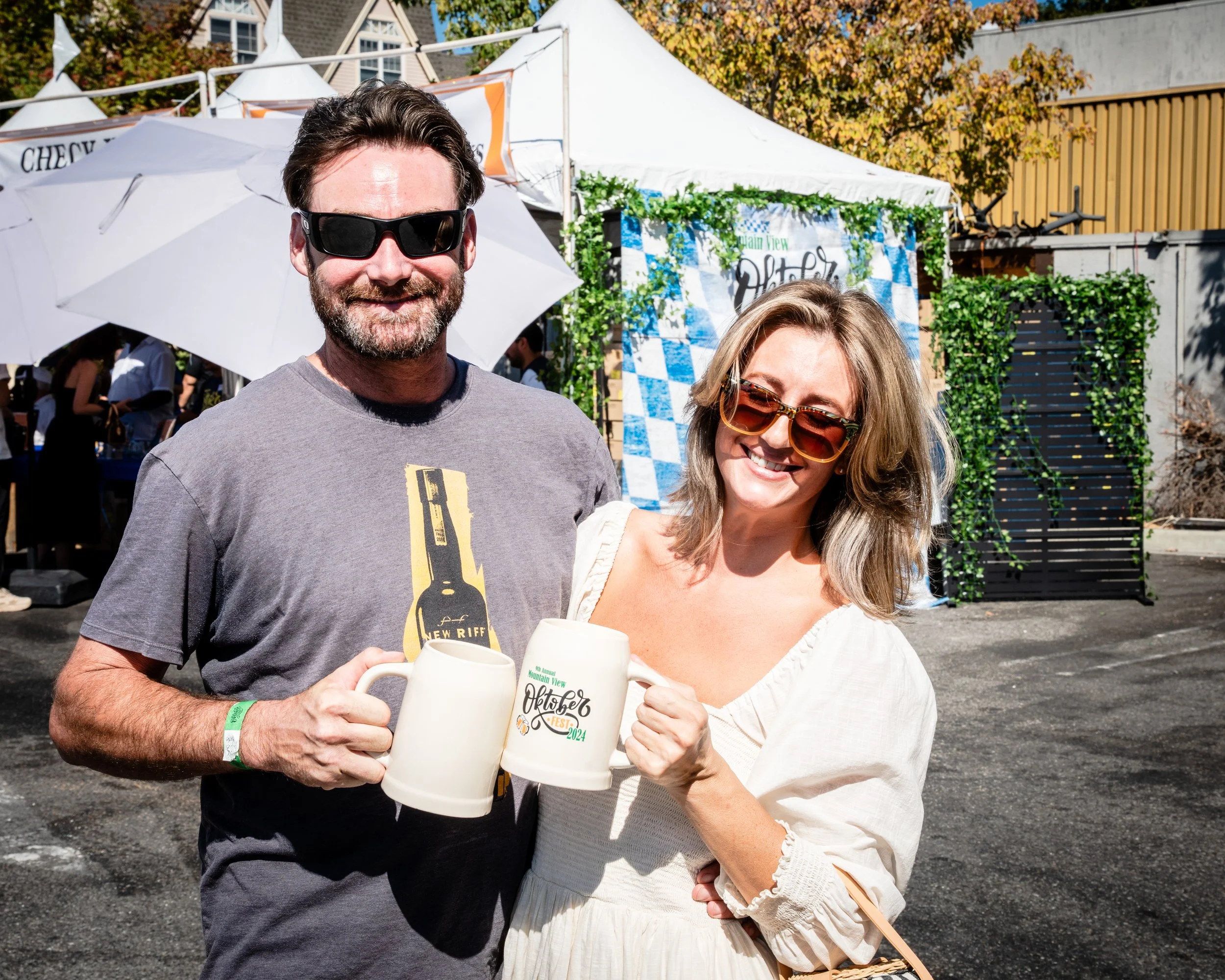 A smiling man and woman are posing outdoors at a festival, each holding a beer stein. The man has a beard and is wearing sunglasses, a grey t-shirt, and a wristband. In the background, there are tents, an umbrella, and decorative greenery with a sign