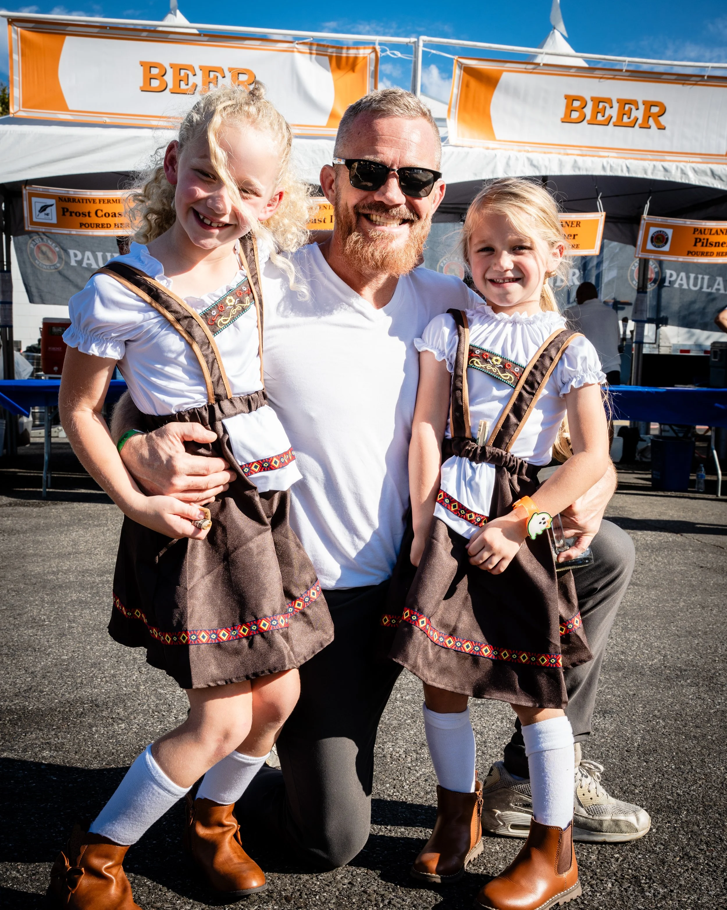 A man with two young girls dressed in traditional Bavarian attire, standing outdoors with booths labeled 'BEER' in the background and smiling at the camera.