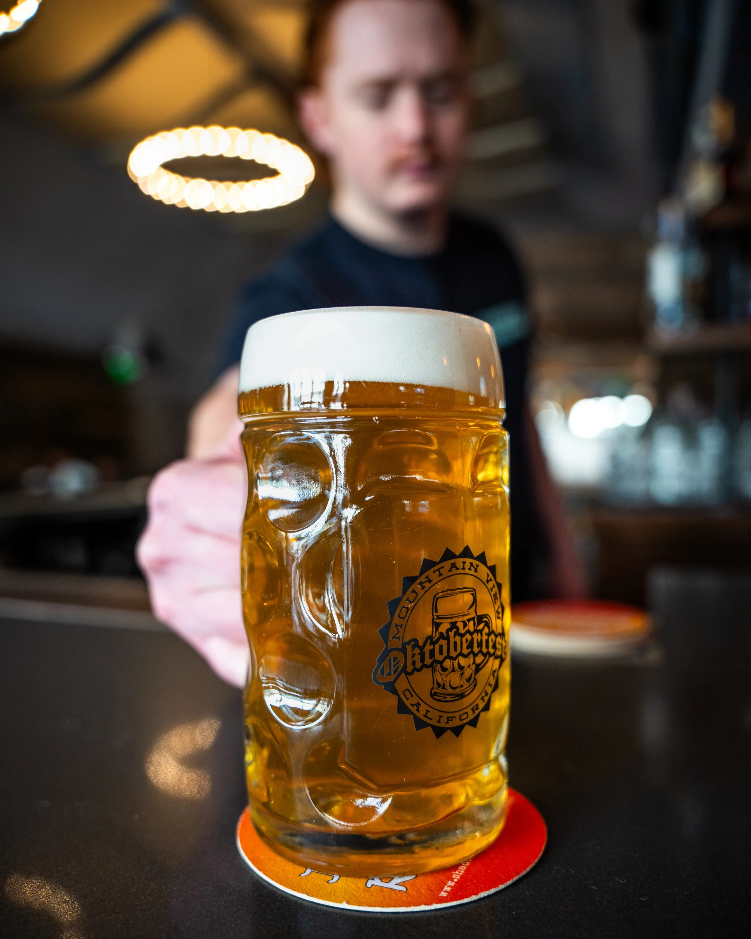 Close-up of a glass of beer with foam on top, held by a person in a bar or pub setting.
