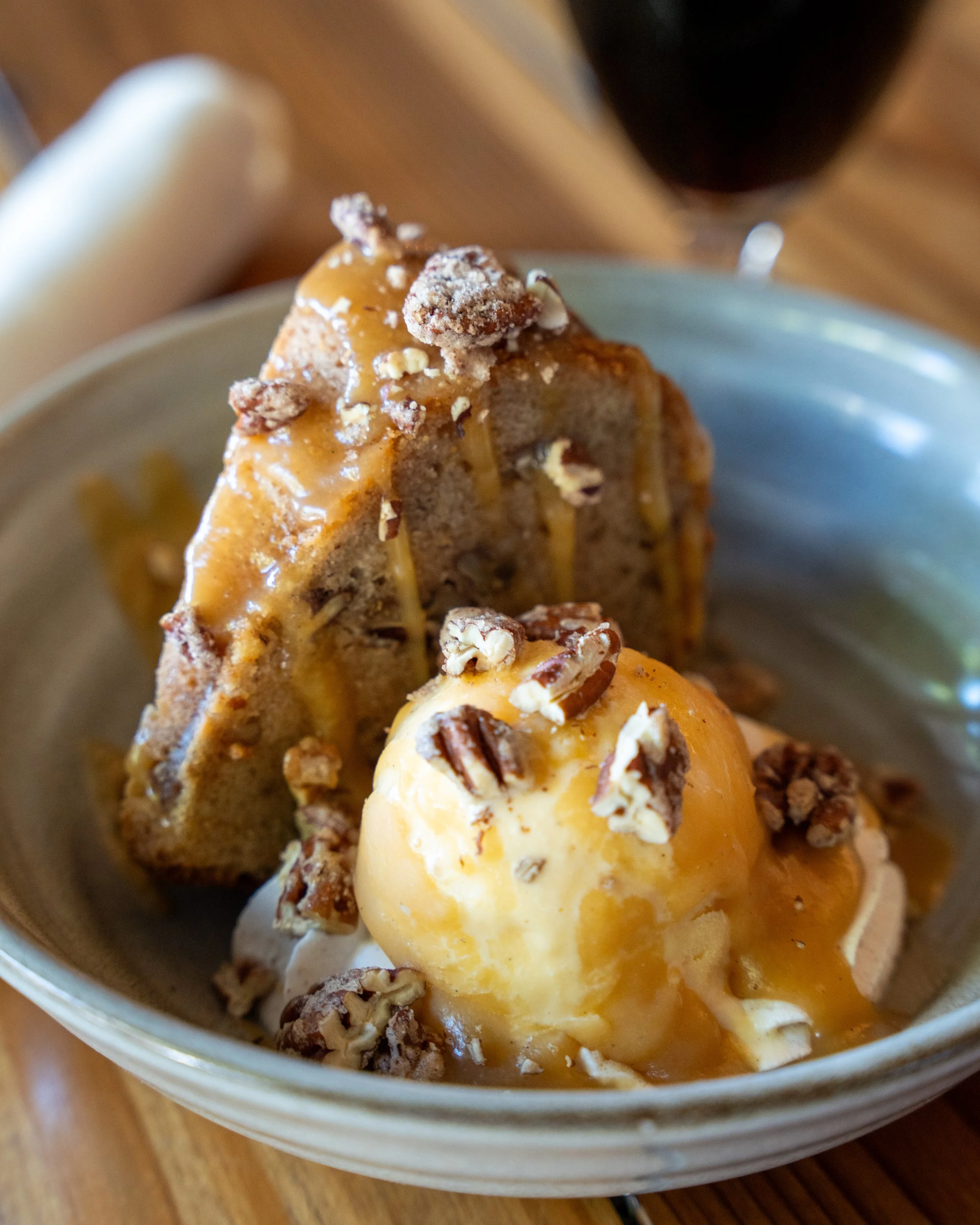 A bowl of bread pudding topped with caramel sauce, chopped pecans, and a scoop of vanilla ice cream.