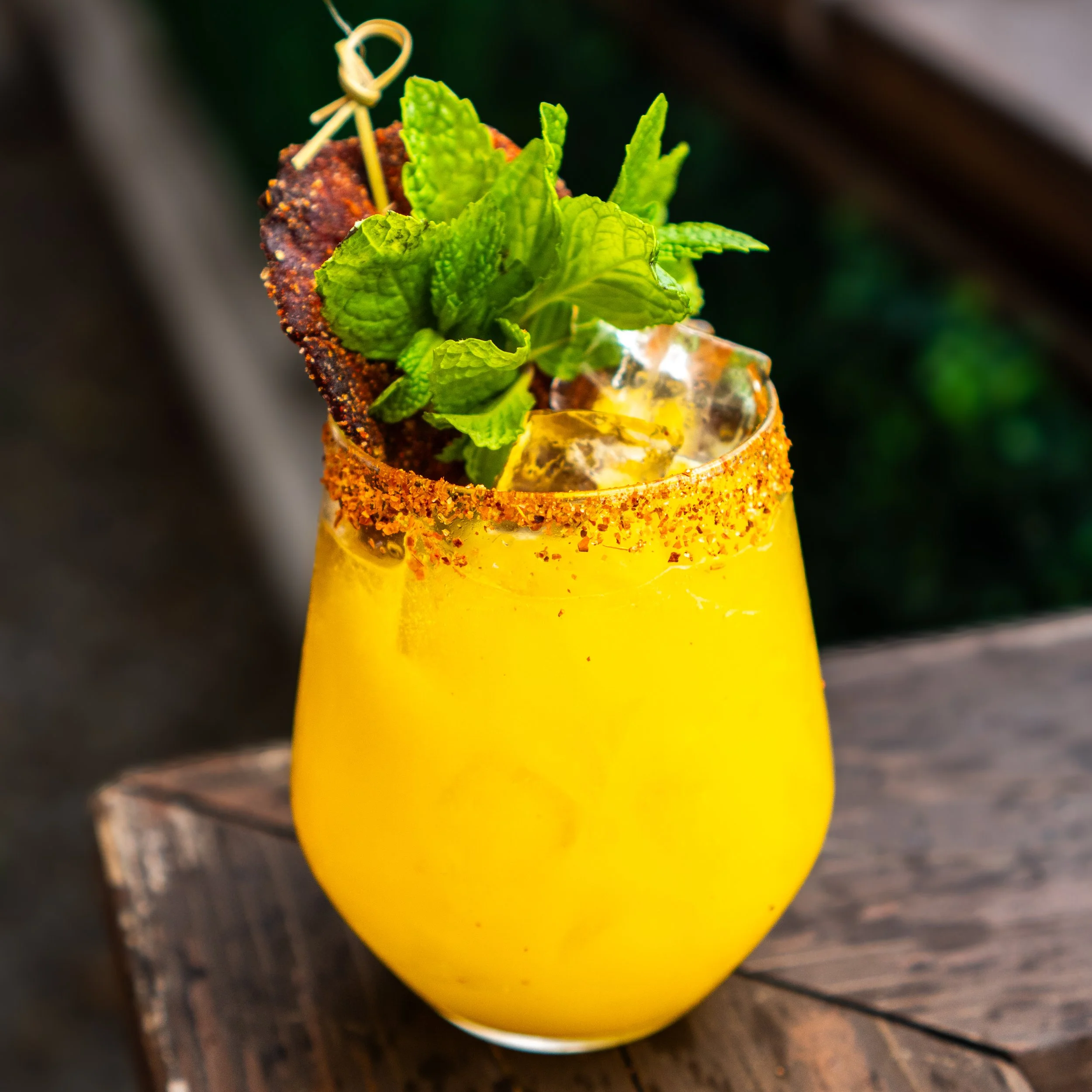 A yellow cocktail garnished with fresh mint, with a chili powder rim and ice, served on a wooden surface.