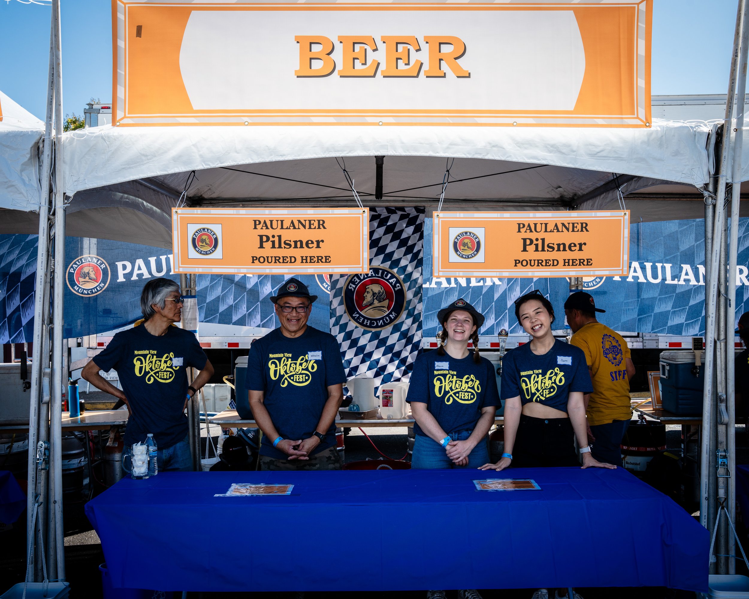 A group of five smiling people standing behind a beer tent at a festival, with signs advertising Paulaner Pilsner and a large orange and white BEER sign. Four of them wear blue shirts with a festival logo, and one wears a yellow staff shirt. The tent