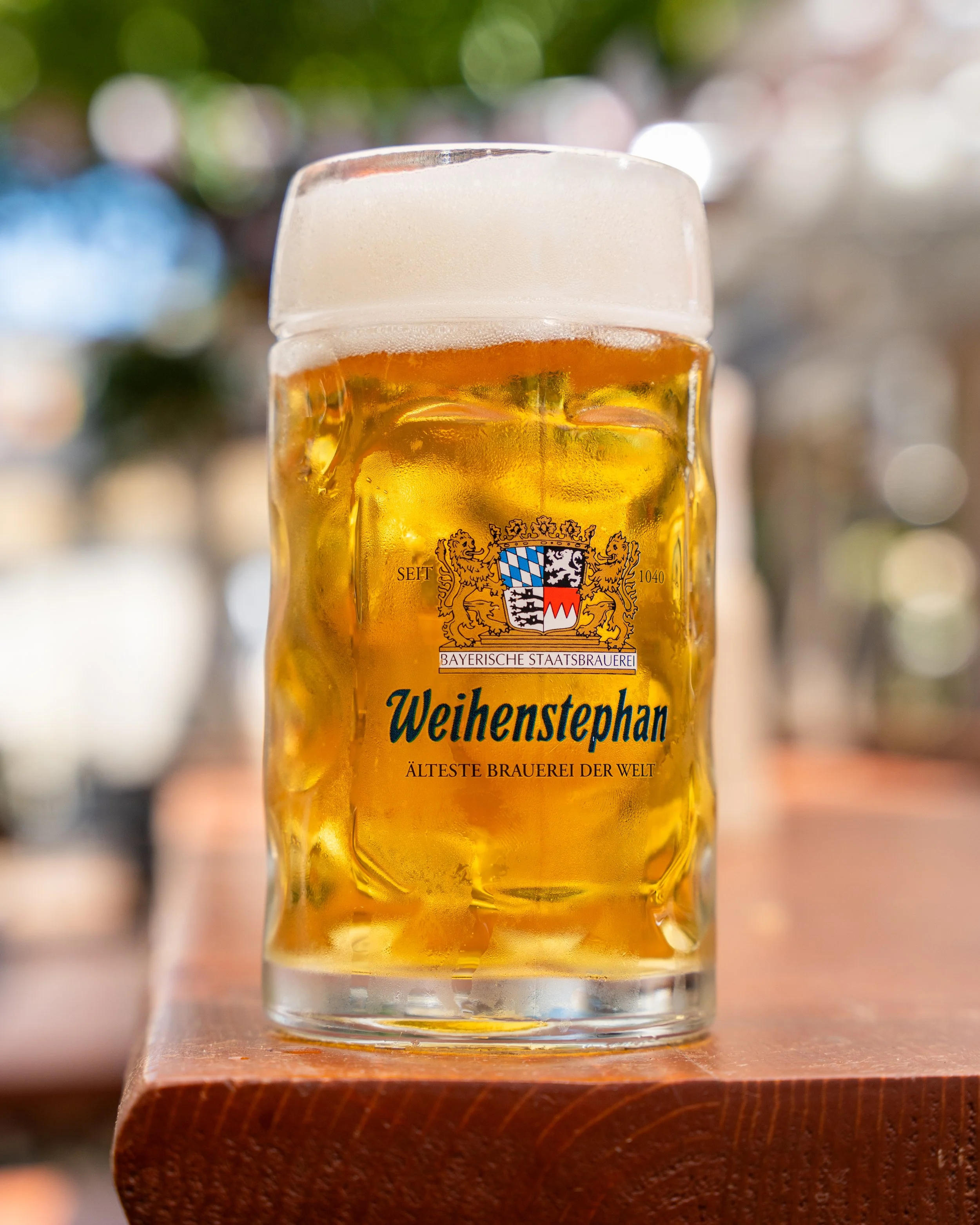 A glass of beer with foam on top, featuring the Weihenstephan brewery logo and coat of arms, set on a wooden surface with a blurred background.