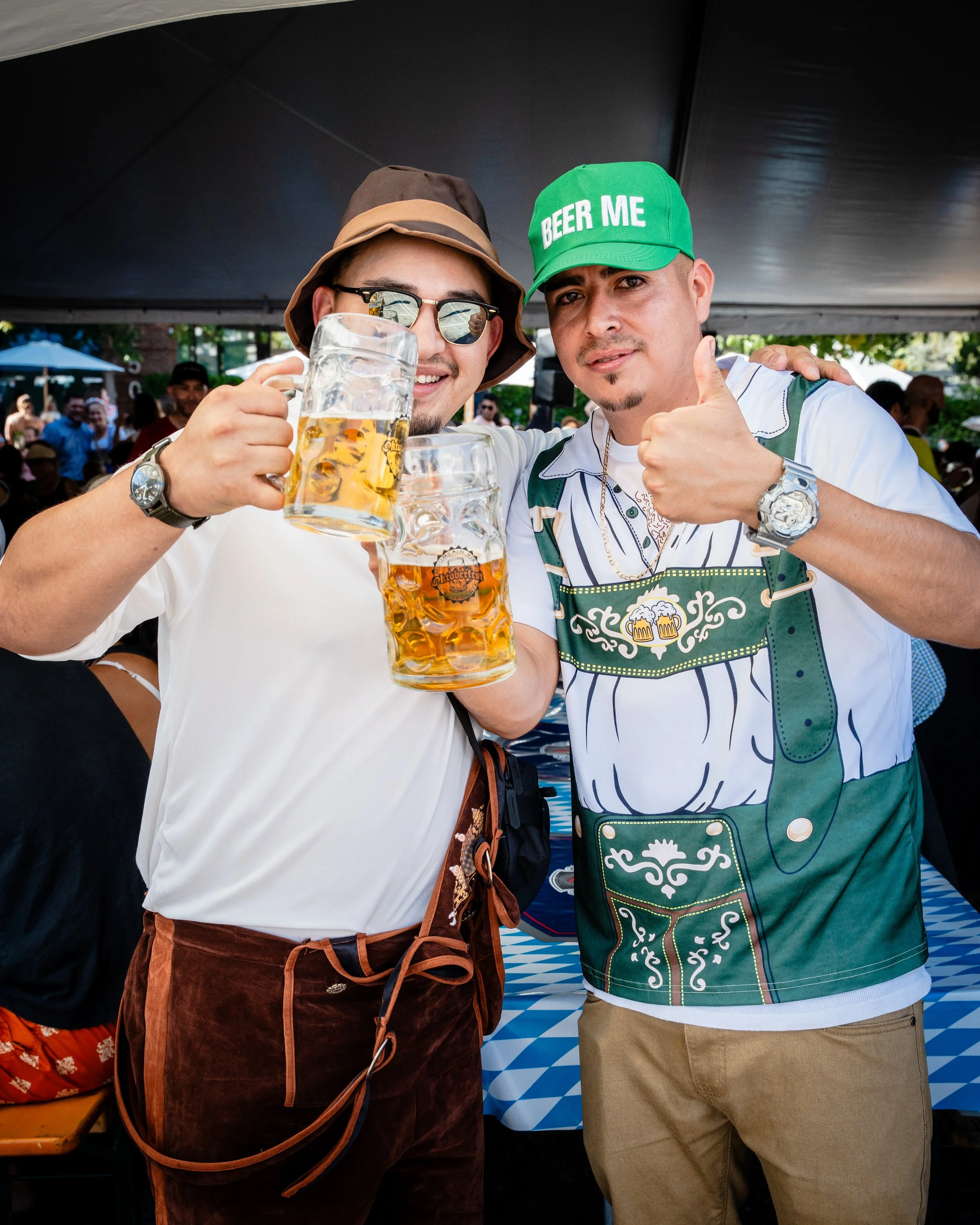 Two men celebrating at an outdoor festival, holding large mugs of beer, wearing casual clothing and accessories, under a tent with a crowd in the background.