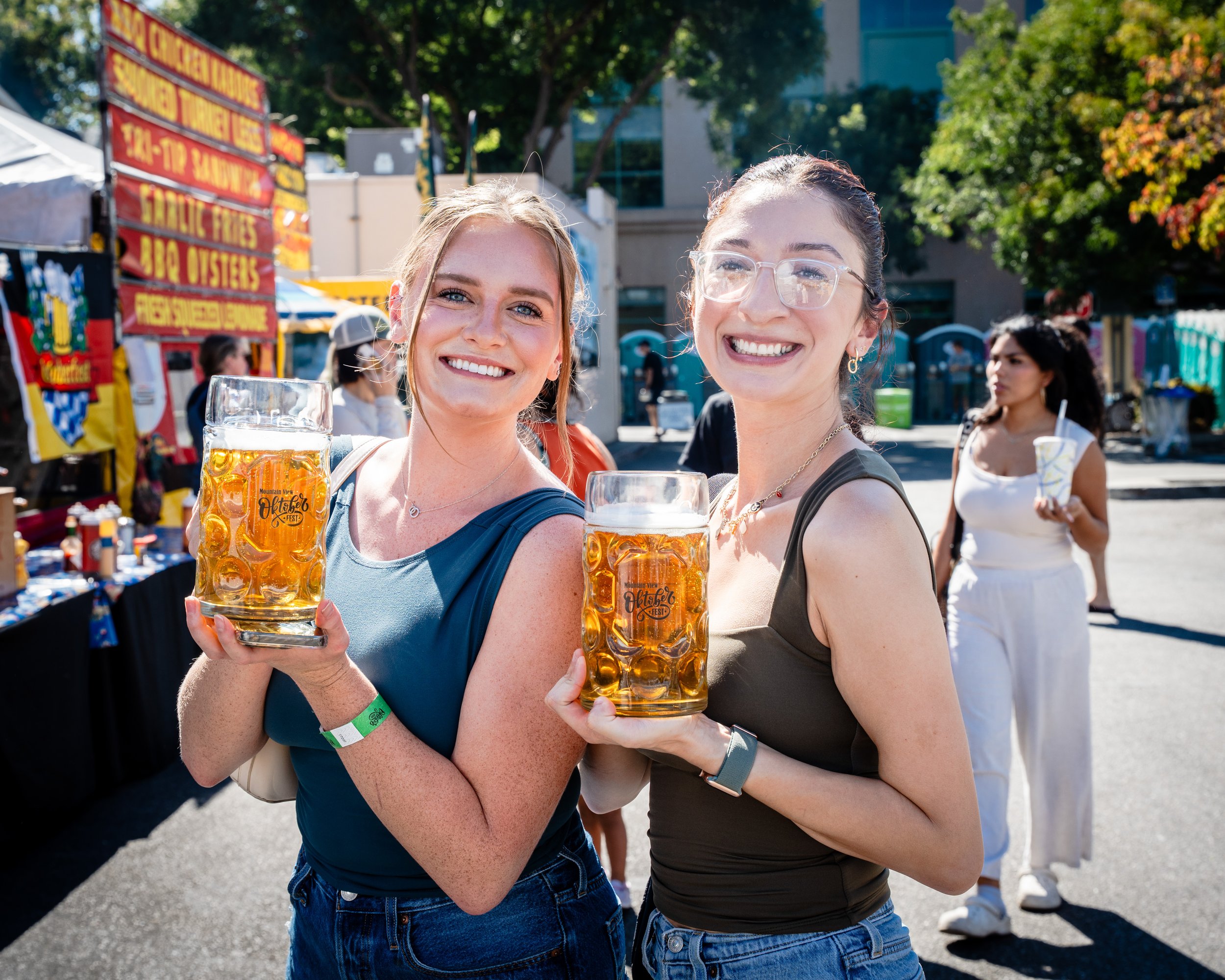 Two women smiling and holding large mugs of beer at an outdoor event or festival on a sunny day, with food and activity tents in the background.