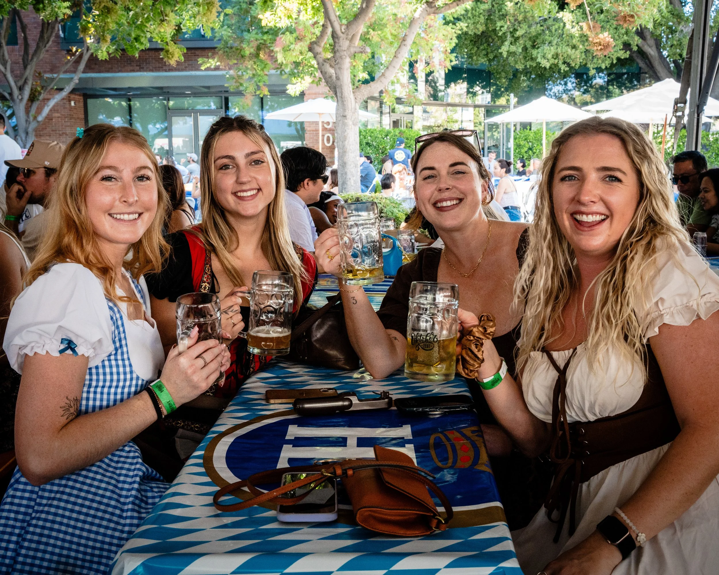 Four women in traditional Bavarian dresses celebrating at an outdoor festival, holding beer mugs, smiling, with other festival-goers and trees in the background.