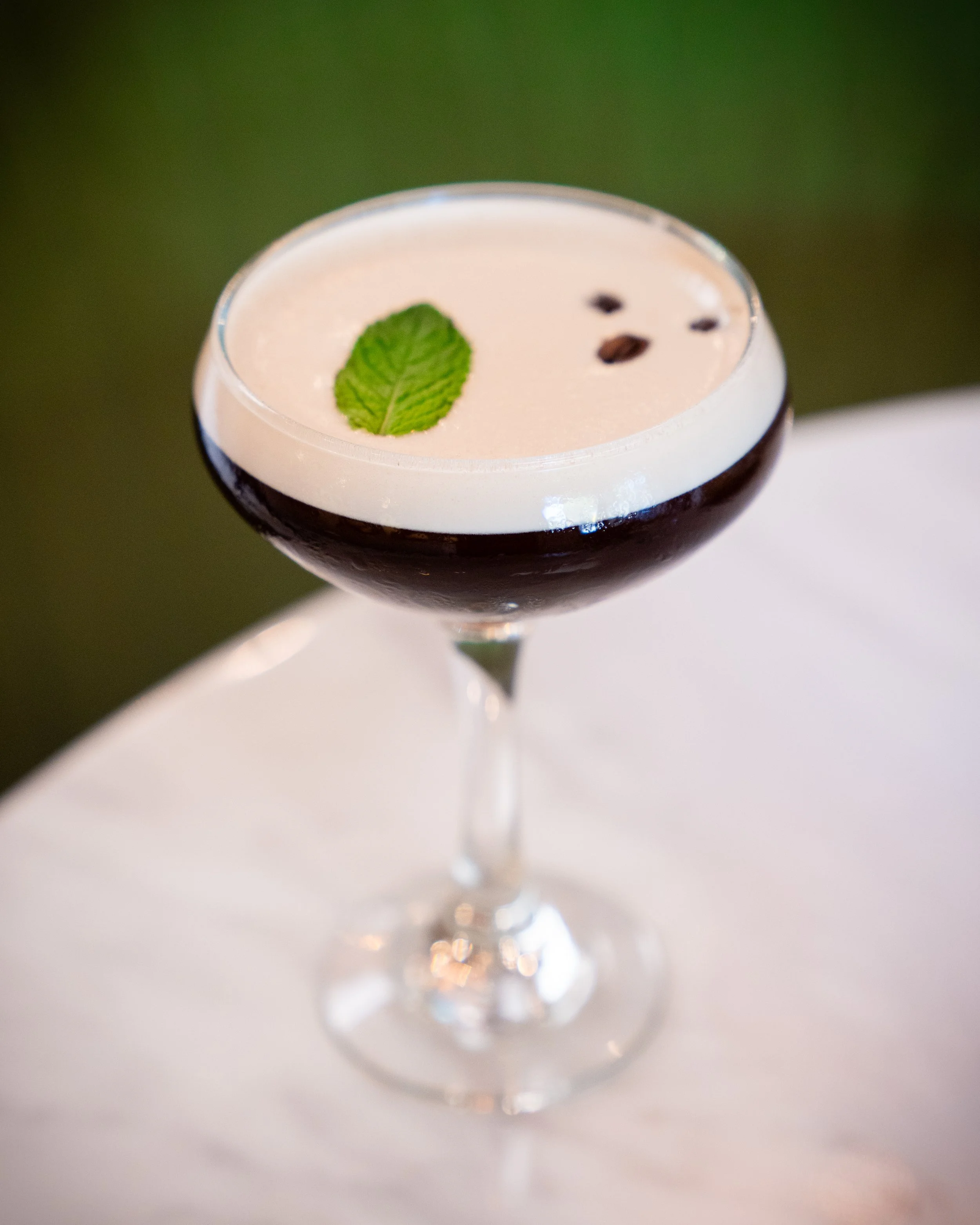A cocktail in a coupe glass with black and white layered drink, garnished with a mint leaf and coffee beans.