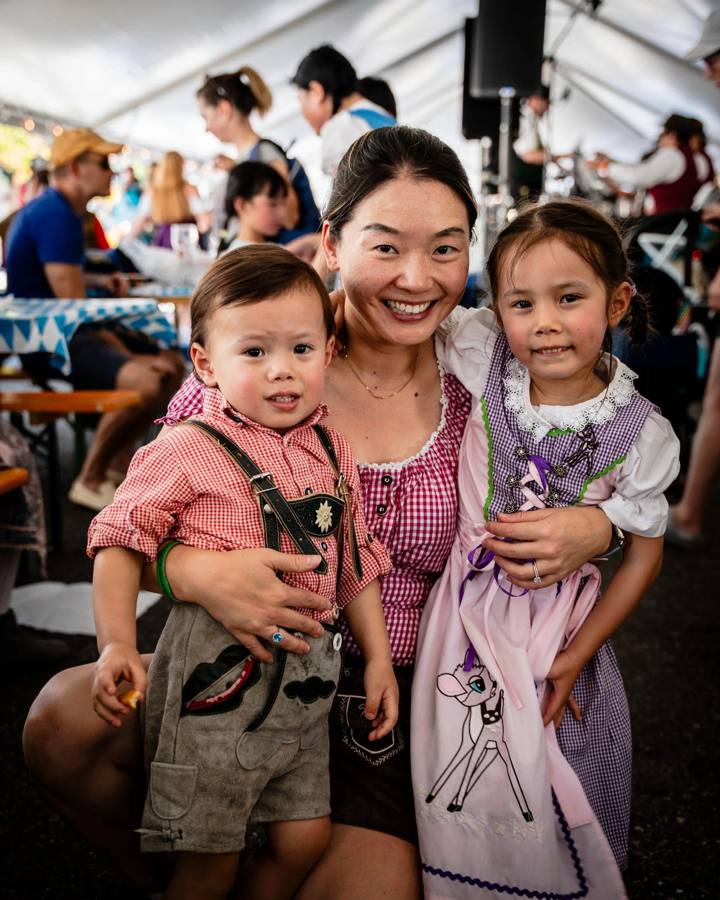 A woman smiling, dressed in a traditional German dirndl, with a young boy in lederhosen and a young girl in traditional dress, at a festive outdoor event under a large white tent.