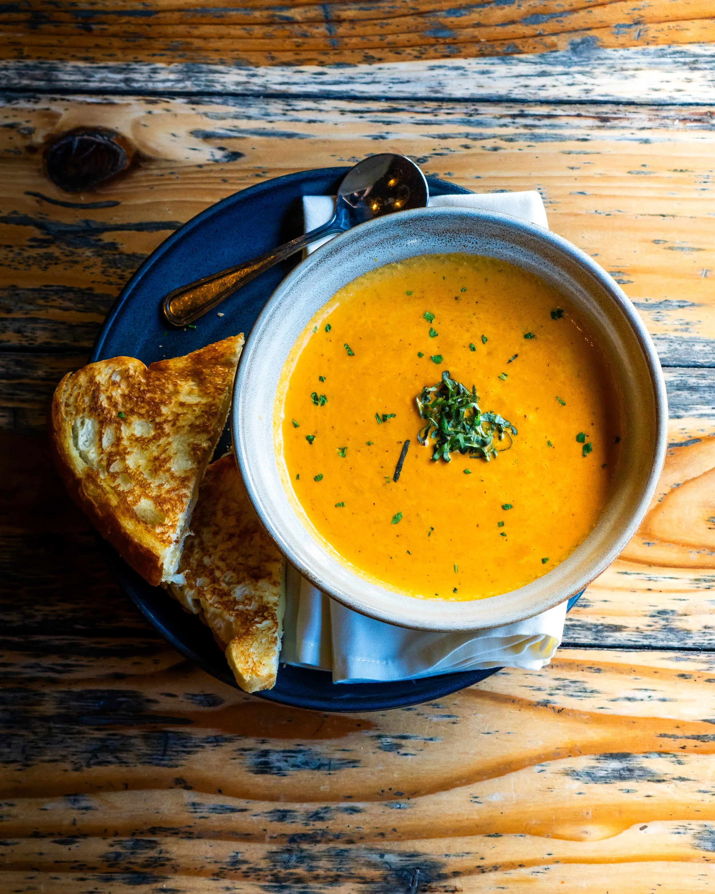 A bowl of creamy orange soup garnished with chopped herbs, accompanied by toasted bread, on a wooden table with a spoon on a napkin.