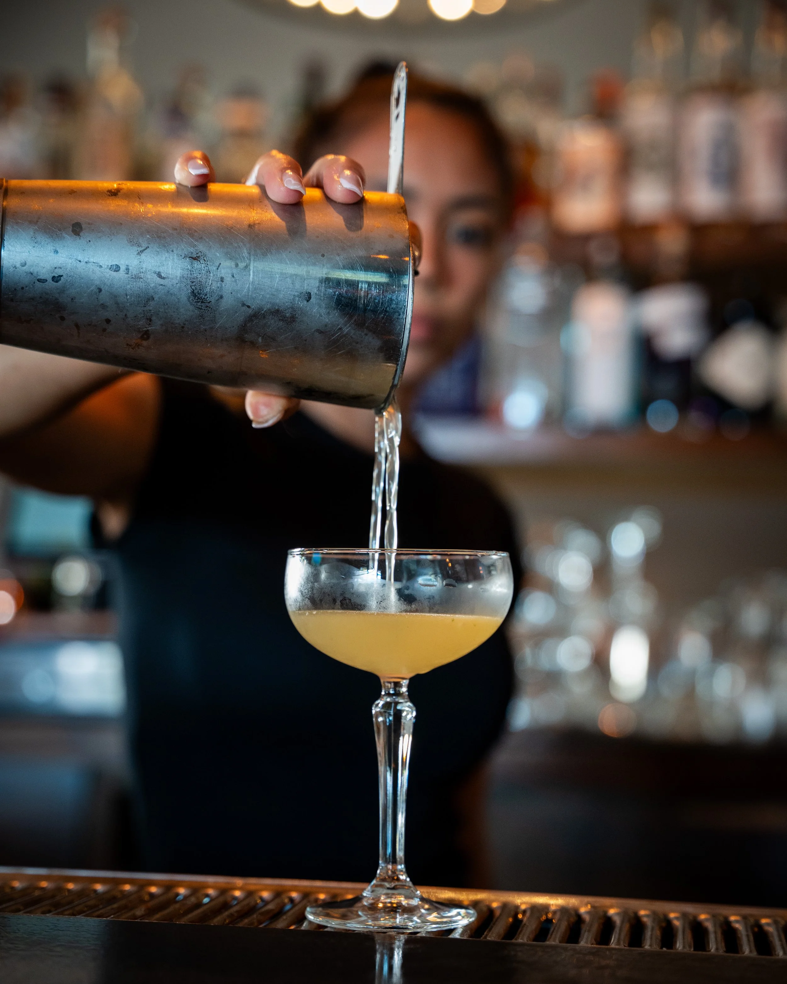 Bartender pouring a yellow cocktail into a glass in a bar setting.