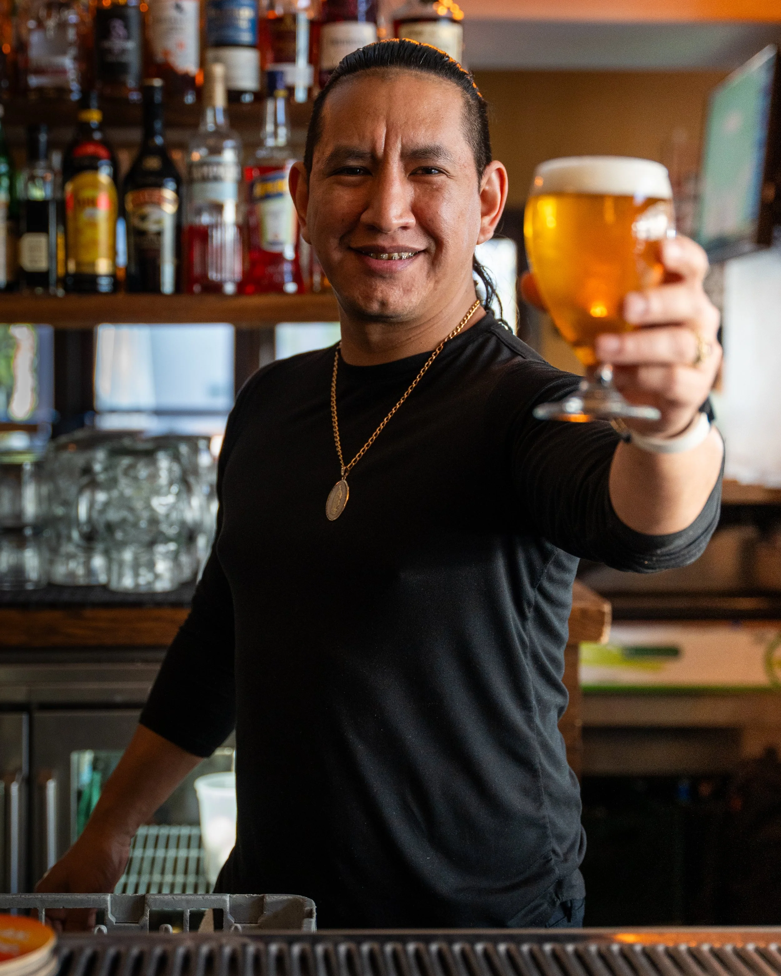 A man with dark hair, wearing a black shirt, gold chain, and ring, is behind a bar counter, holding up a glass of beer and smiling at the camera. The background shows bottles of alcohol on shelves.