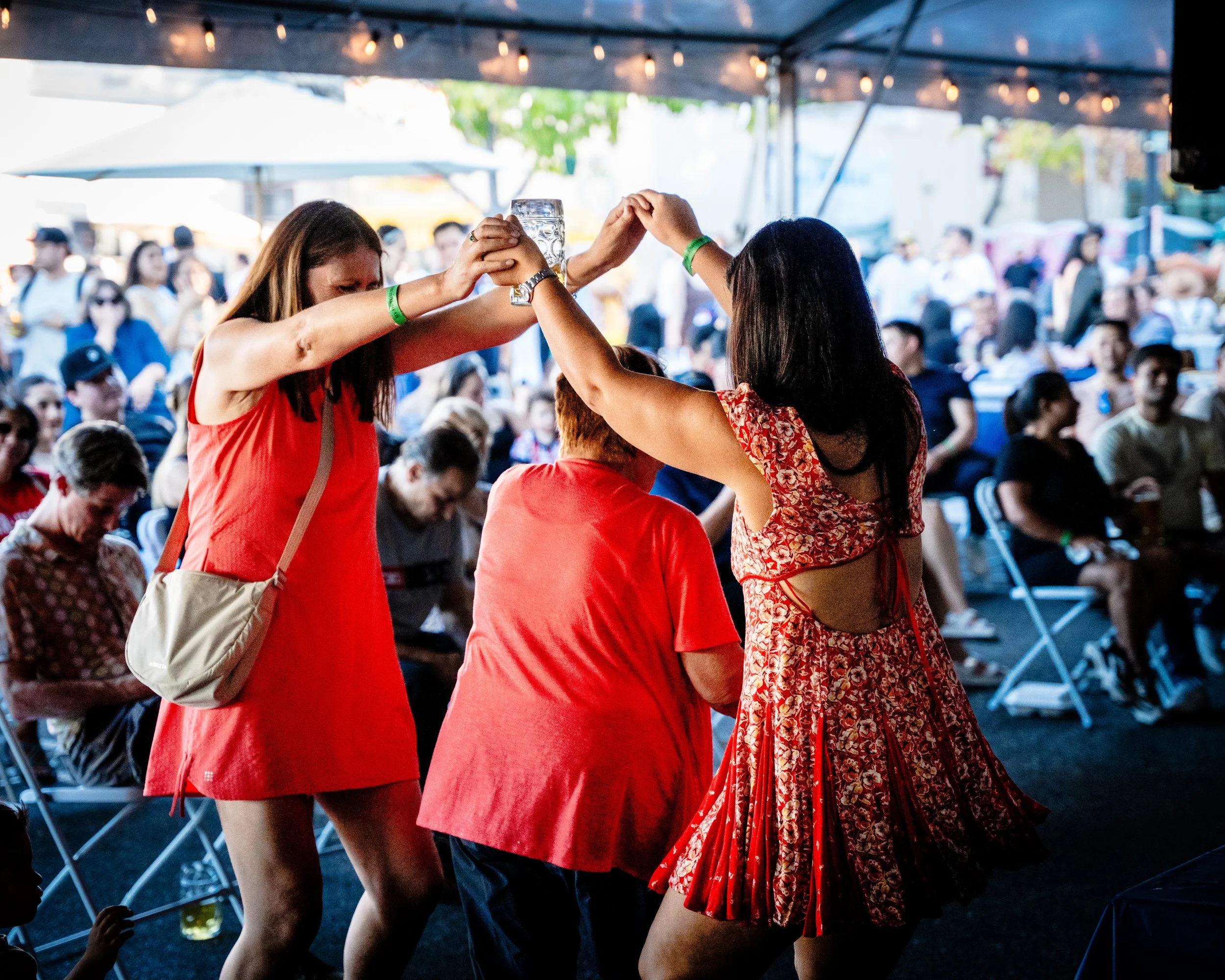 Three women dancing at an outdoor event under a tent, with people seated in the background.