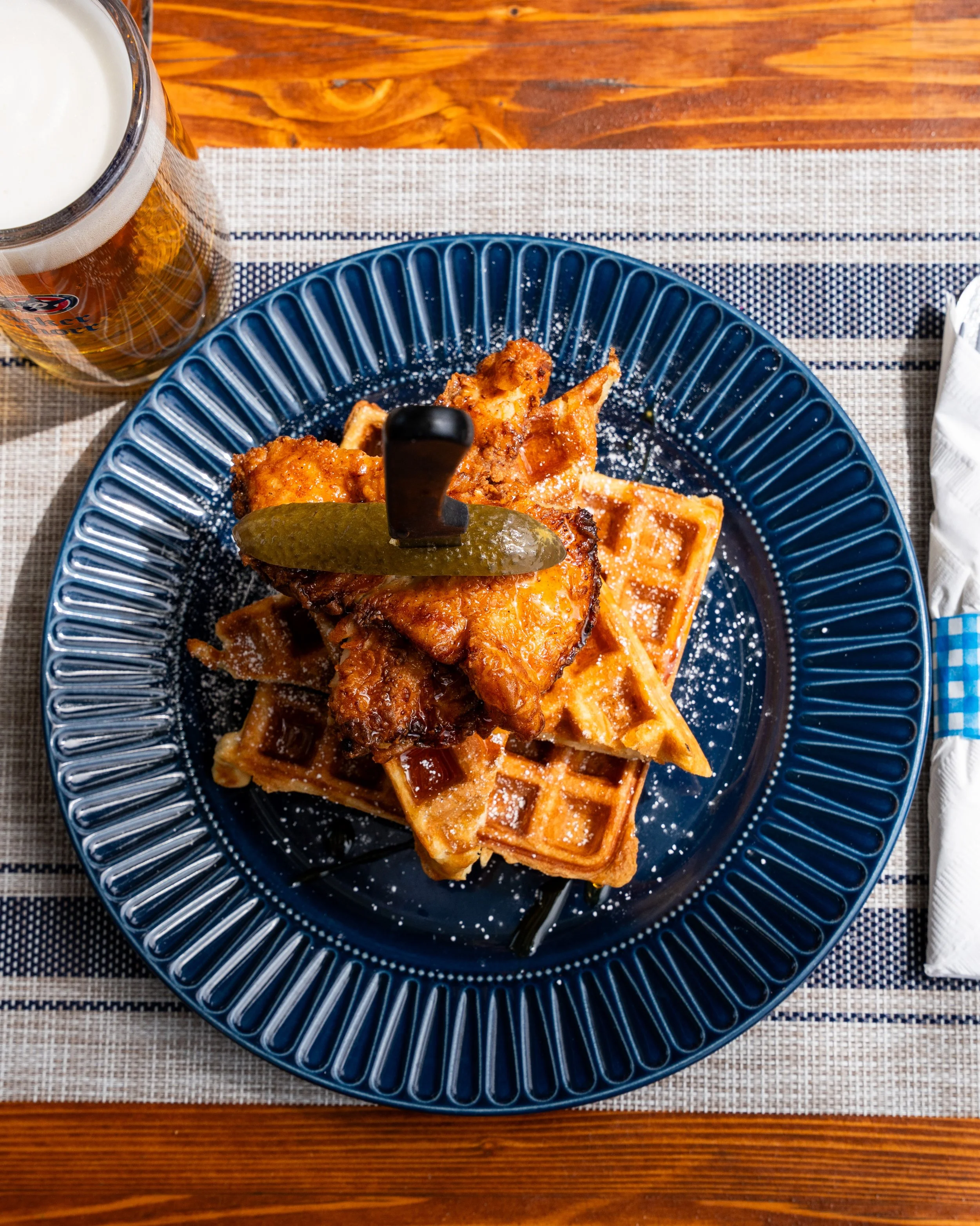 A plate of Belgian waffles topped with fried chicken, a pickle spear, and a small jar of syrup. There is a glass of beer to the left of the plate on a striped tablecloth.