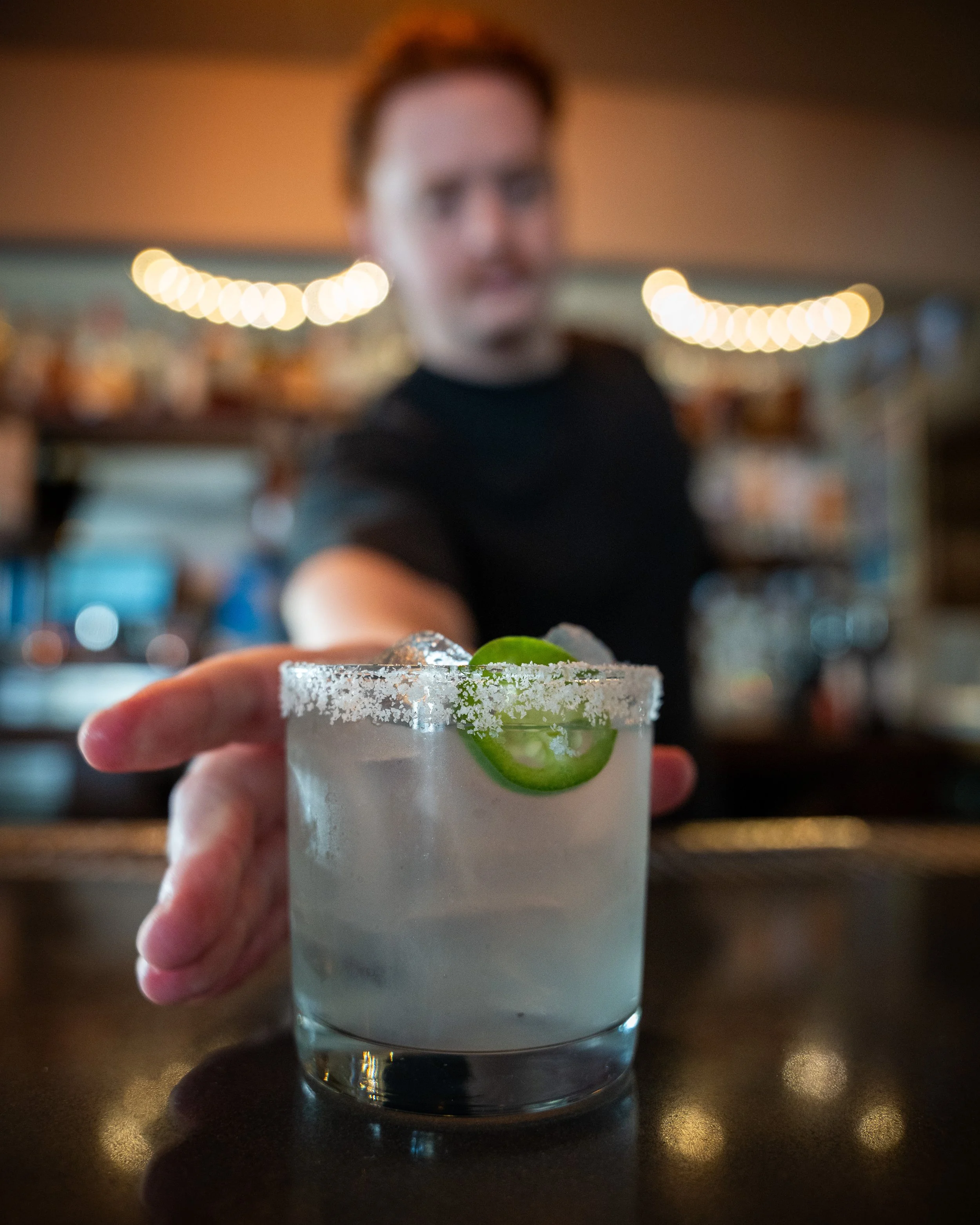 A person behind a bar presenting a cocktail glass with a salted rim, lime slices, and garnishes, out of focus.