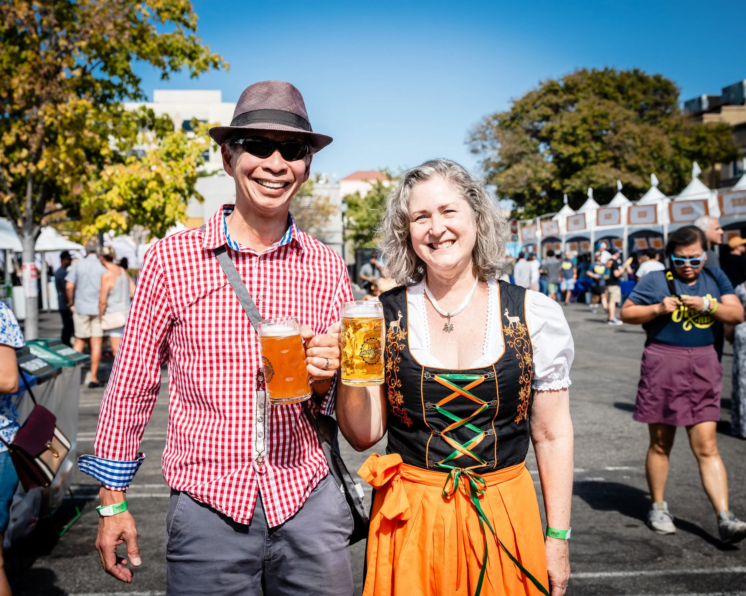 A man and woman dressed in traditional and festive outfits, smiling and holding beer mugs at an outdoor event or festival under a clear blue sky.