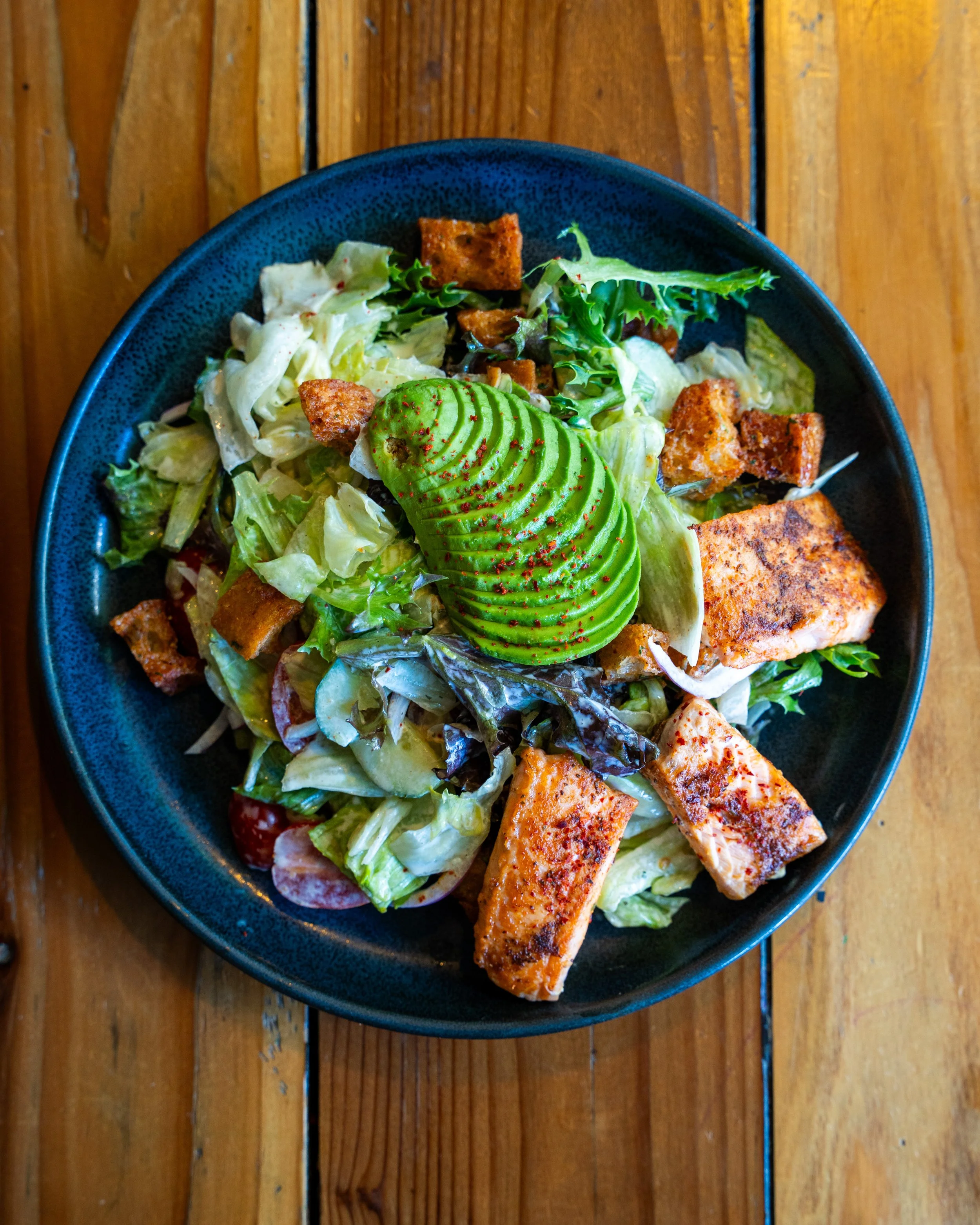 Salad with various greens, grilled salmon pieces, avocado slices, croutons, and red seasoning, served in a blue bowl on a wooden table.