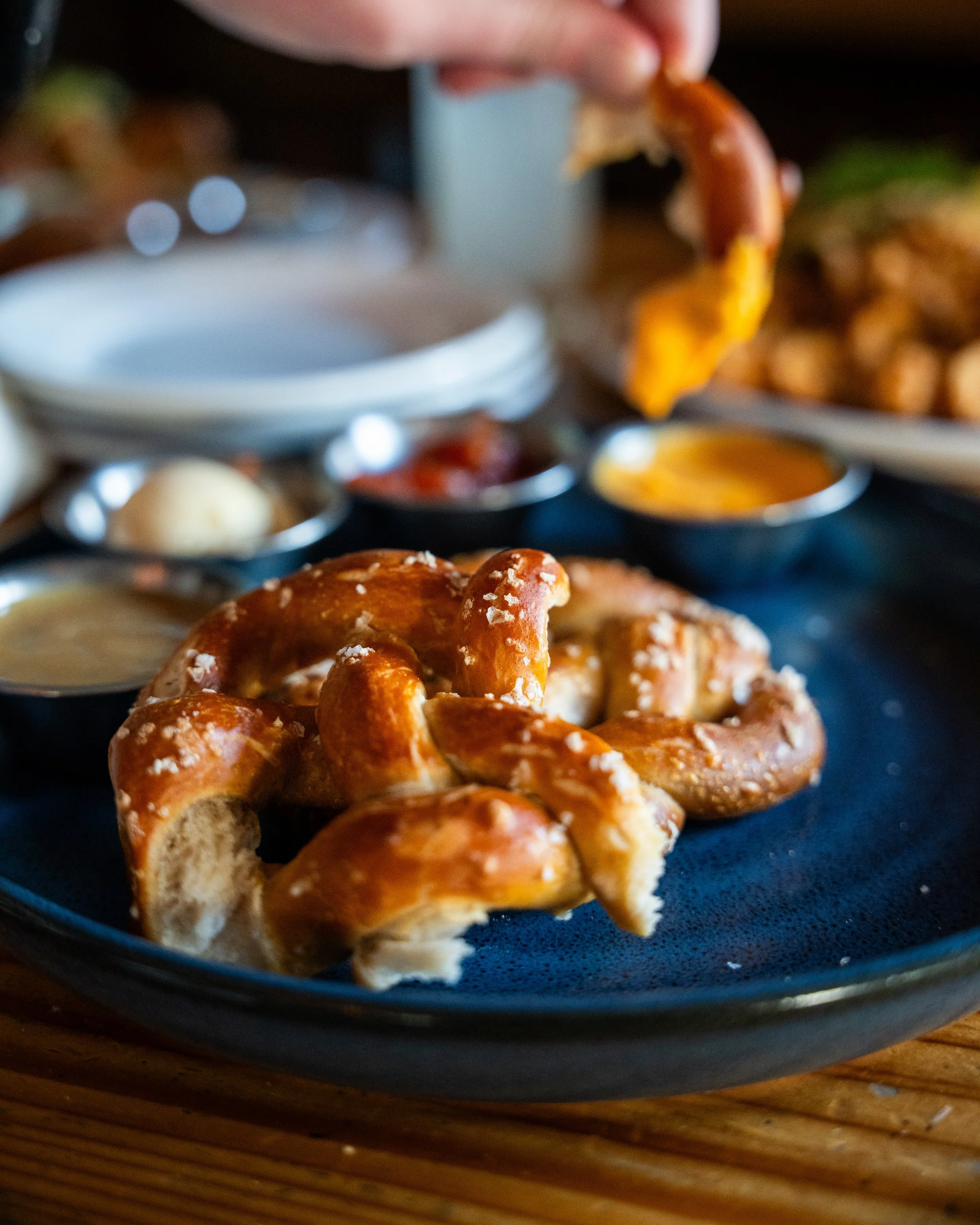 Pretzel with coarse salt on black plate, with dipping sauces and appetizers in background.