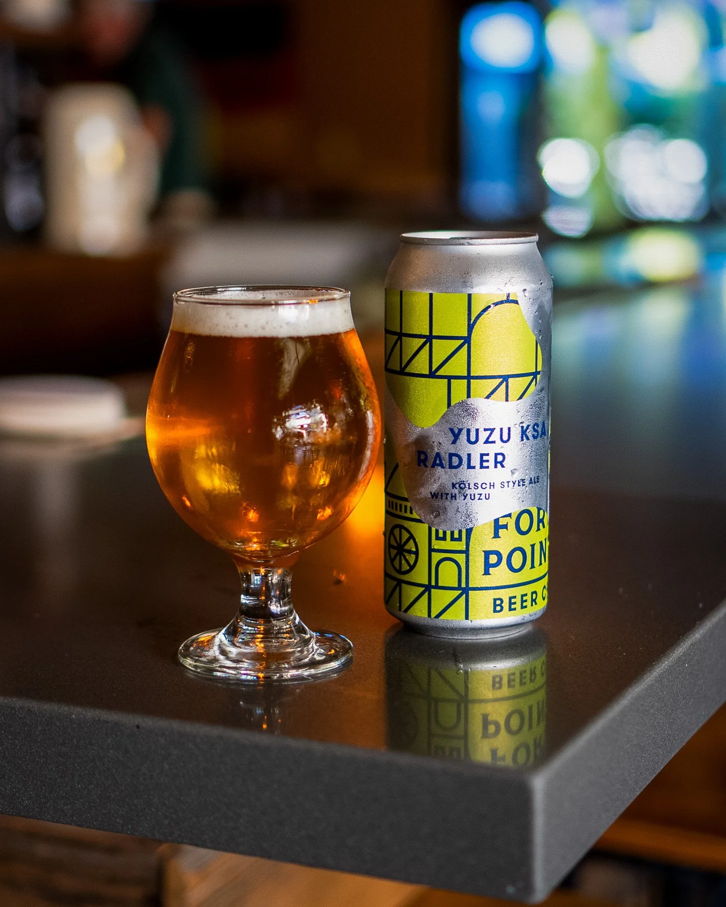 A glass of amber-colored beer and a can of Yuzu Ksa Radler on a dark table with a blurred background.