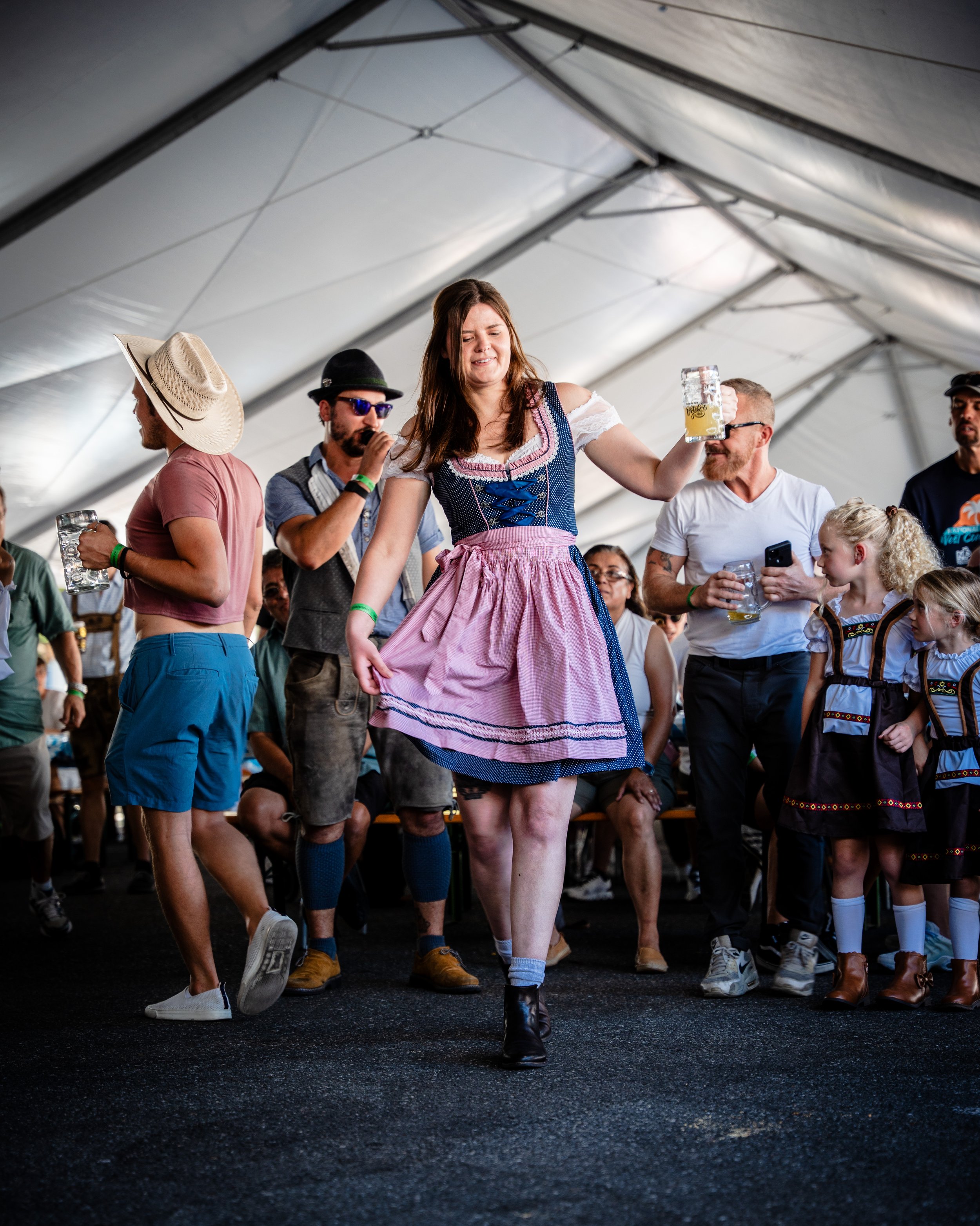 A woman dressed in a traditional dress dancing with a beer mug in a festival tent, surrounded by people also wearing traditional and casual clothing, some holding beer mugs and others using a phone, attending a lively event.
