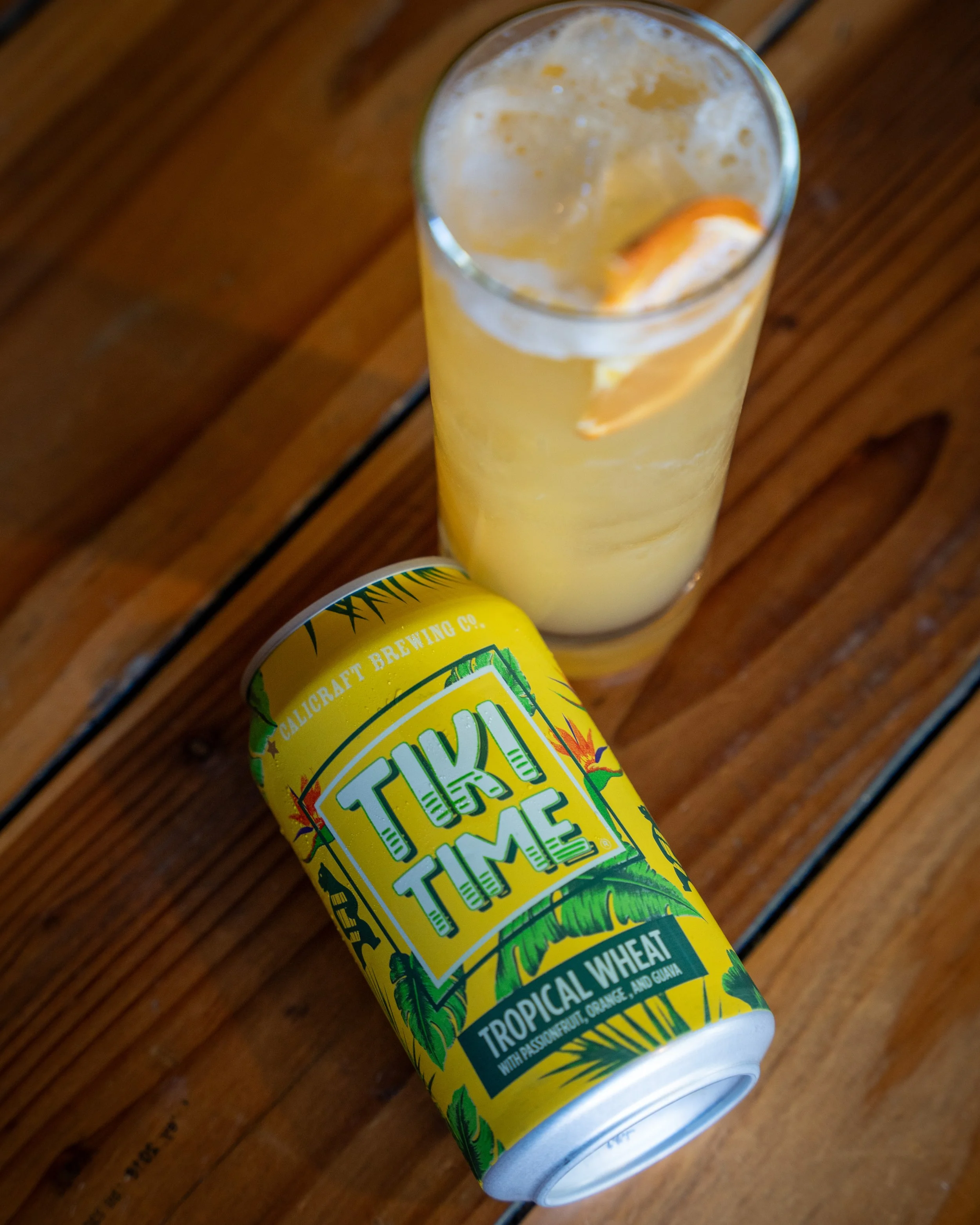 A can of Tuk Tuks Tropical Wheat beer next to a tall glass of beer with a lemon wedge on a wooden table.