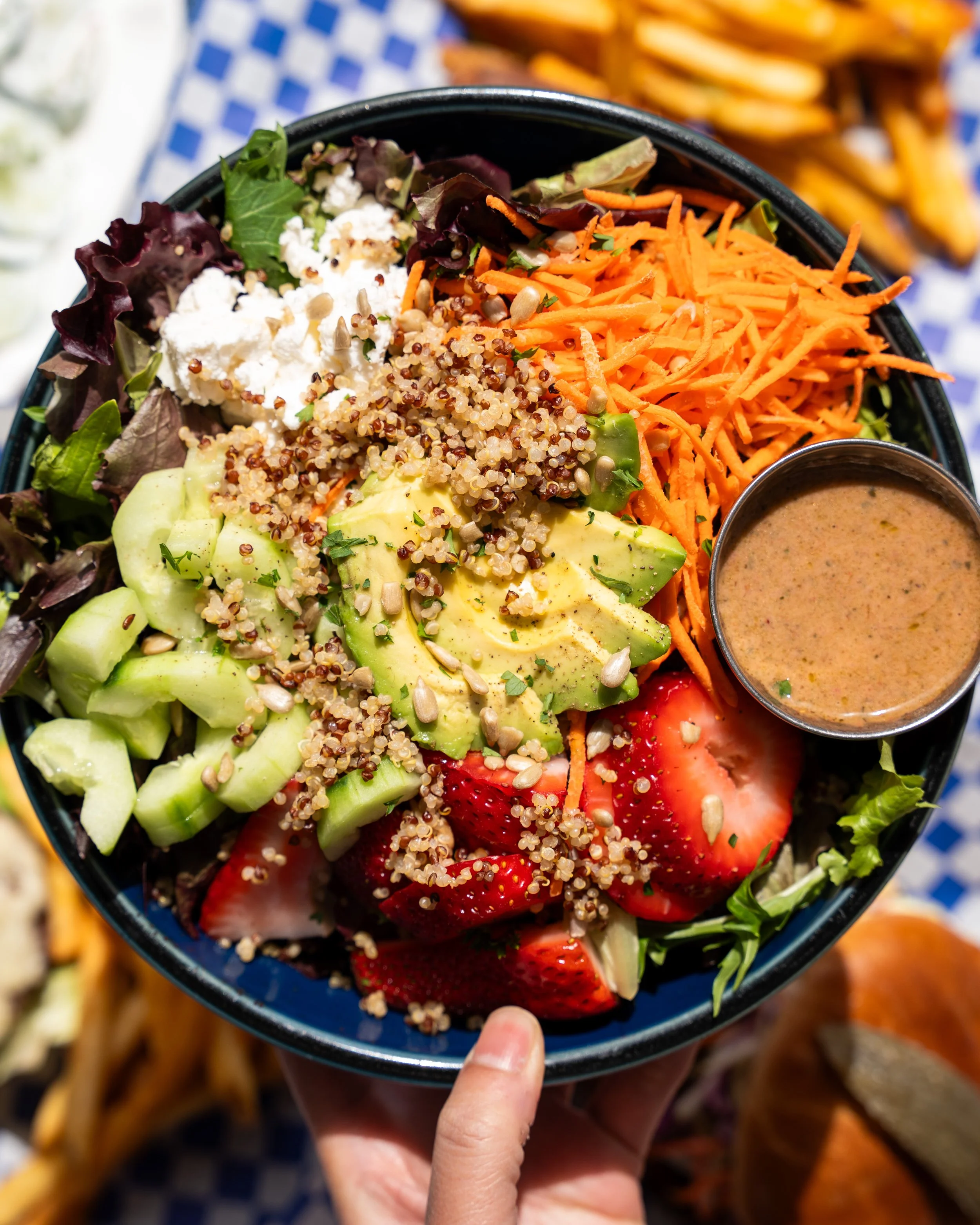 A colorful mixed salad with greens, cherry tomatoes, avocado, shredded carrots, quinoa, crumbled cheese, sunflower seeds, and a side of dressing in a small metal cup.