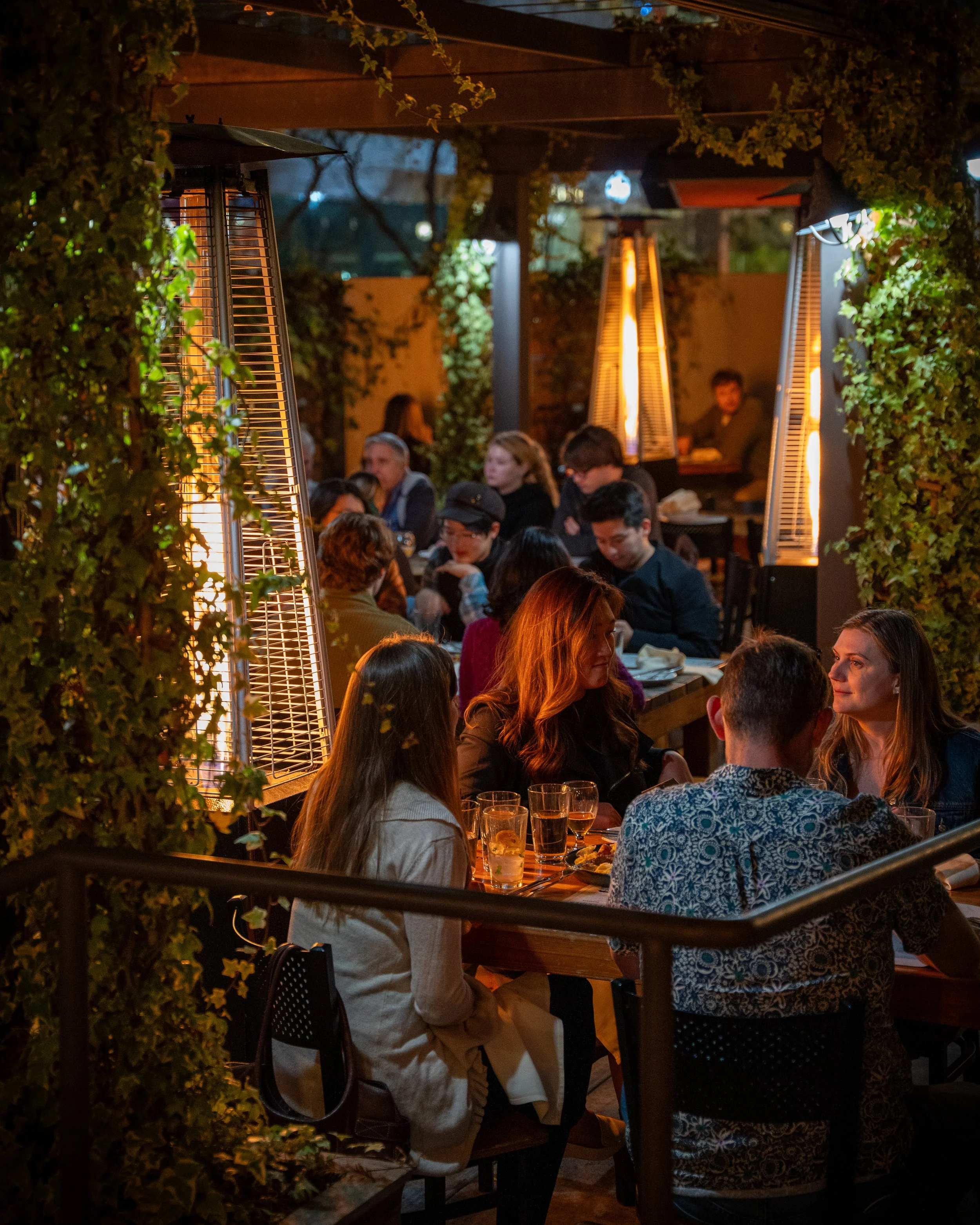People dining outdoors at a restaurant during evening with warm lighting and heaters, surrounded by greenery.