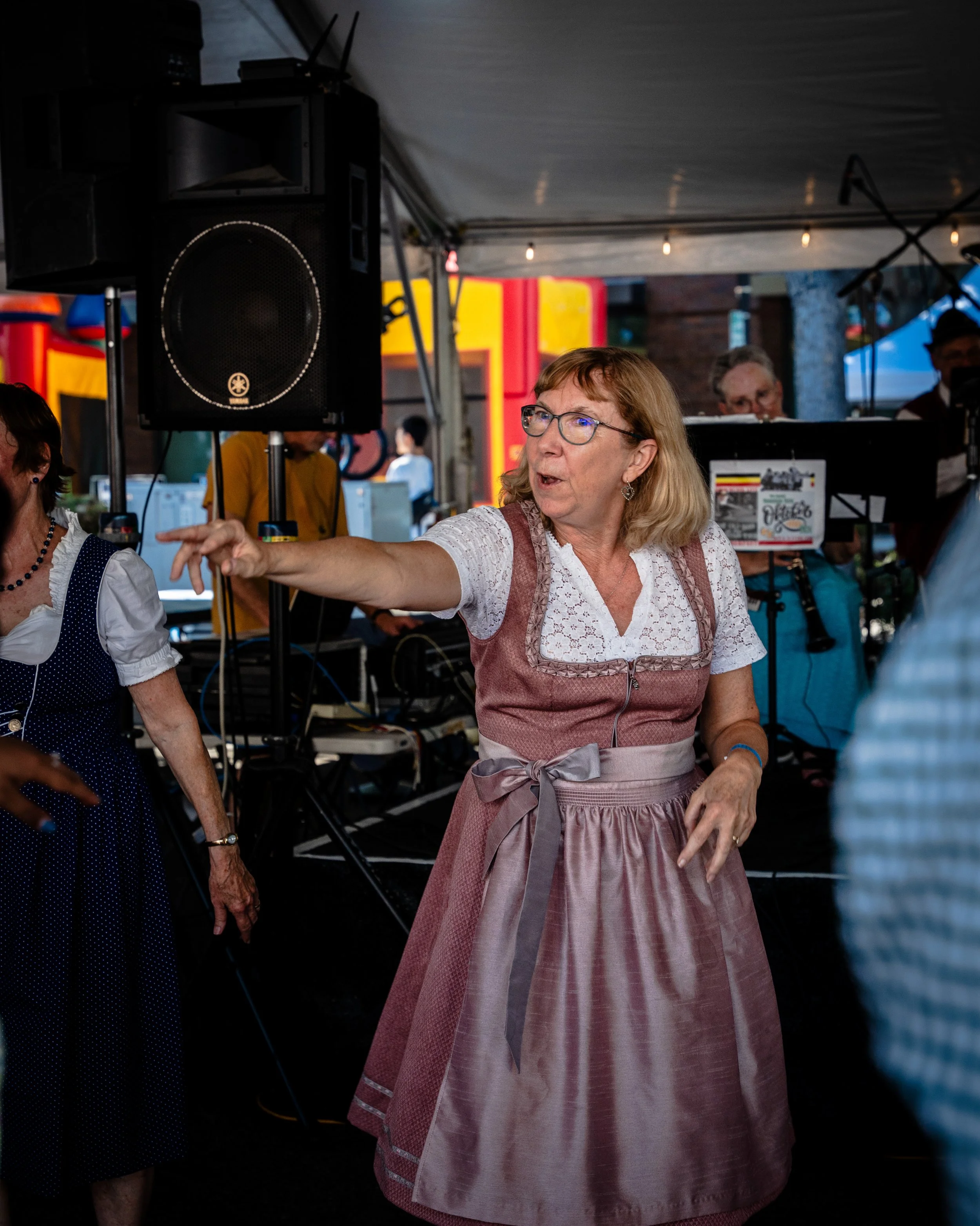 A woman wearing a traditional dress with a pink apron and glasses is pointing while talking at an event under a tent, with a band visible in the background and a speaker nearby.