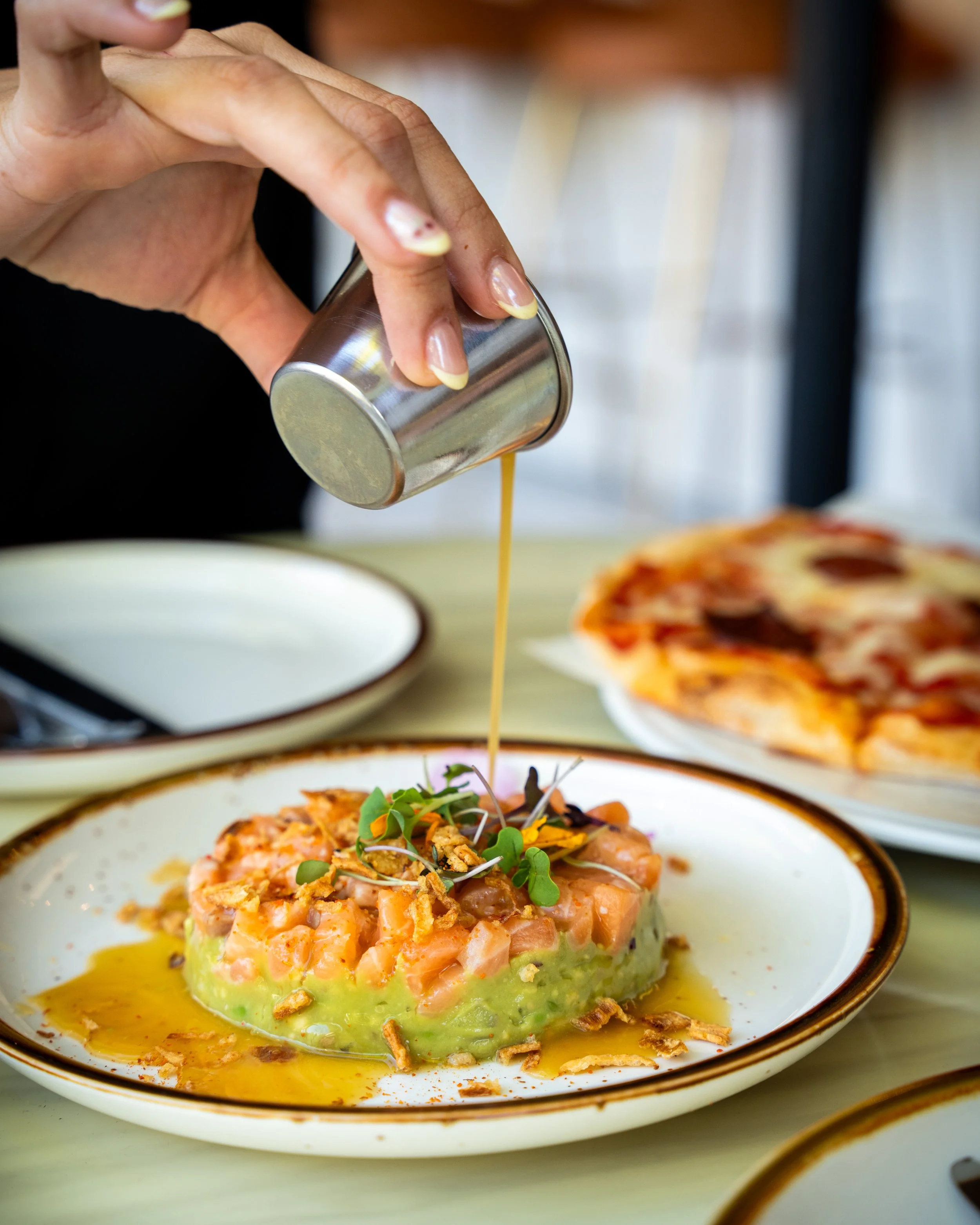 Person pouring dressing over a dish of diced salmon, avocado, and microgreens on a white plate at a restaurant table, with a pepperoni pizza in the background.