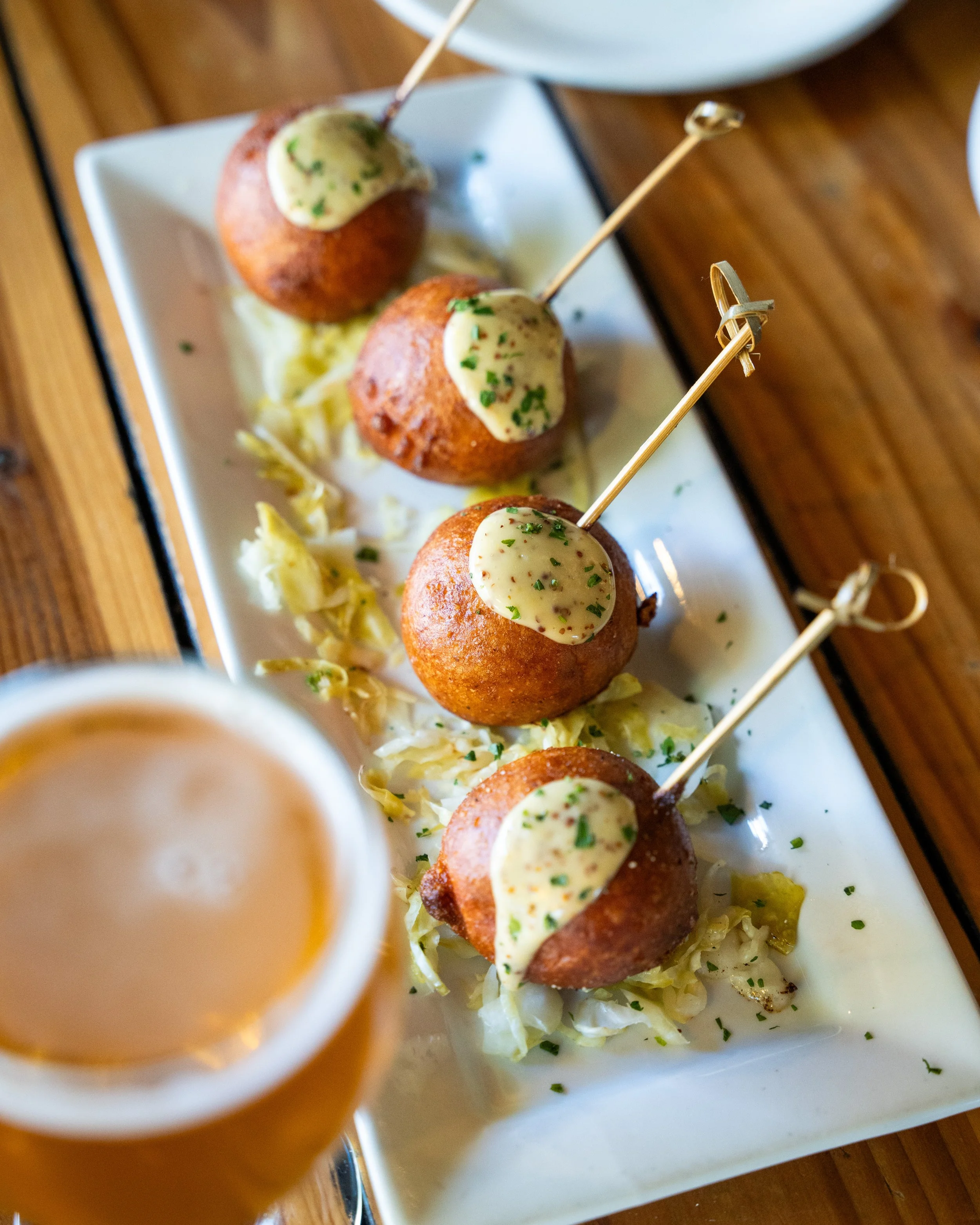 Four fried ball appetizers topped with creamy sauce and chopped herbs, served on a white plate with shredded lettuce and a glass of beer in the foreground.