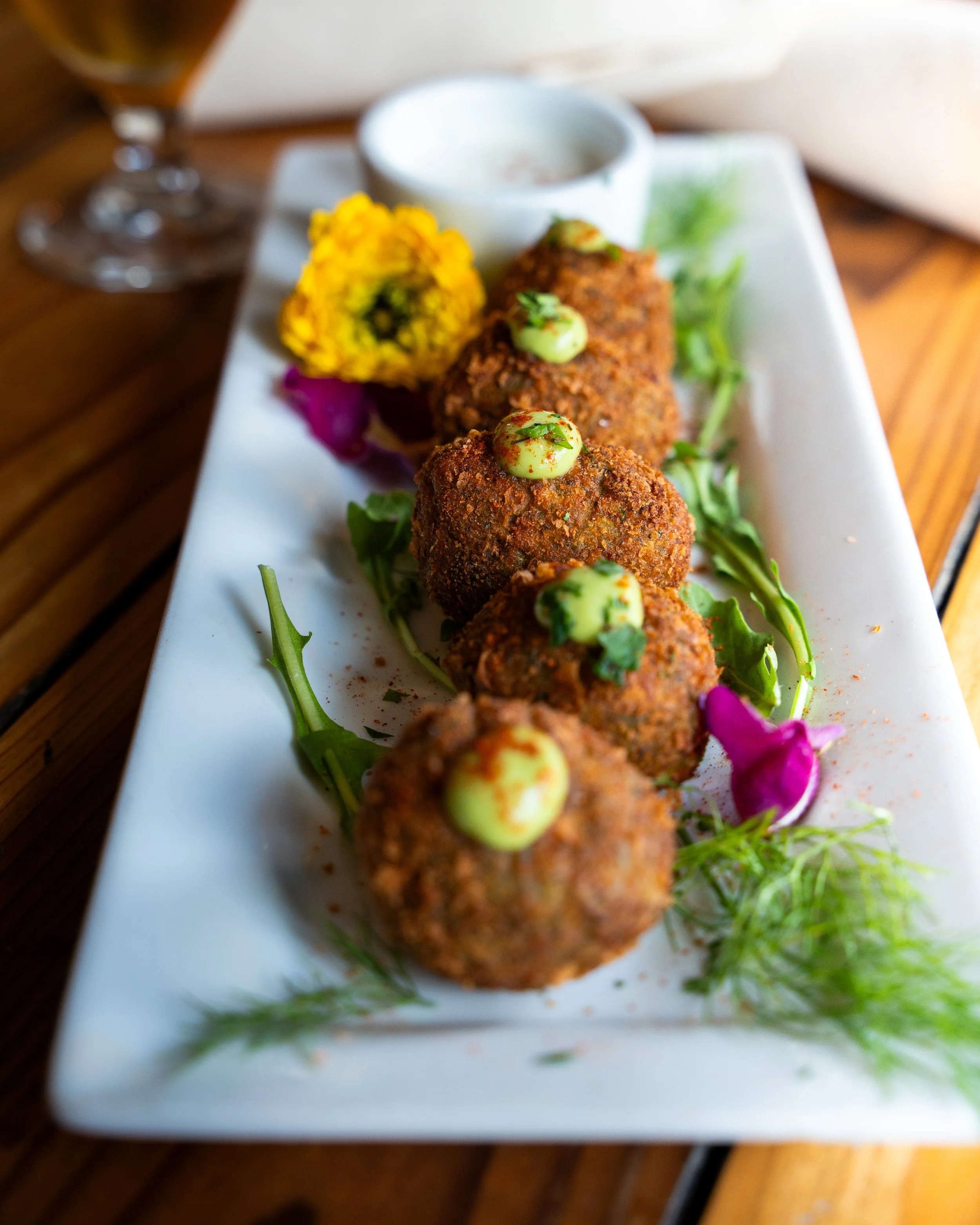 Fried appetizer balls garnished with green sauce, served with edible flowers and greens on a rectangular white plate.