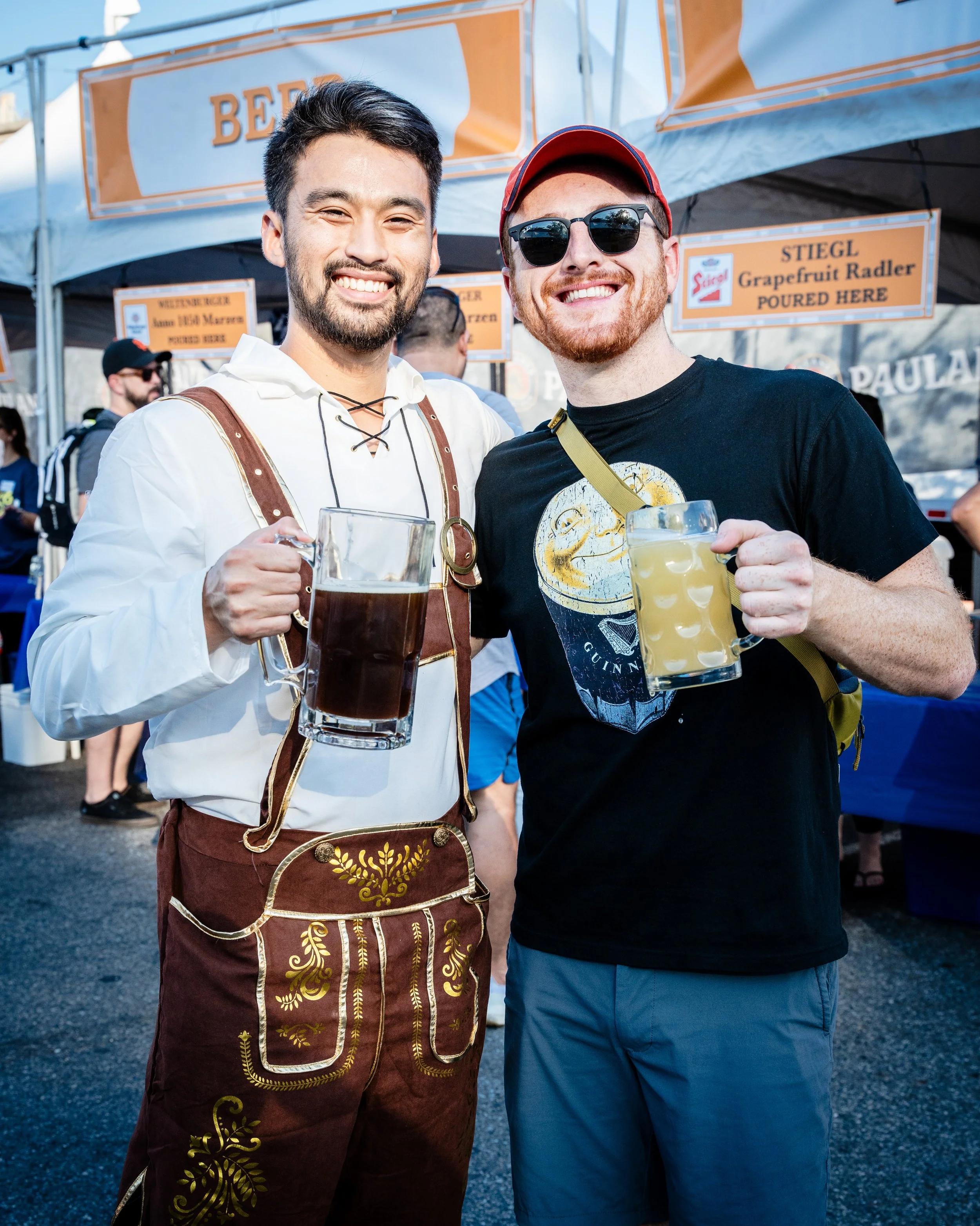 Two men smiling and holding mugs of beer at an outdoor festival or event.