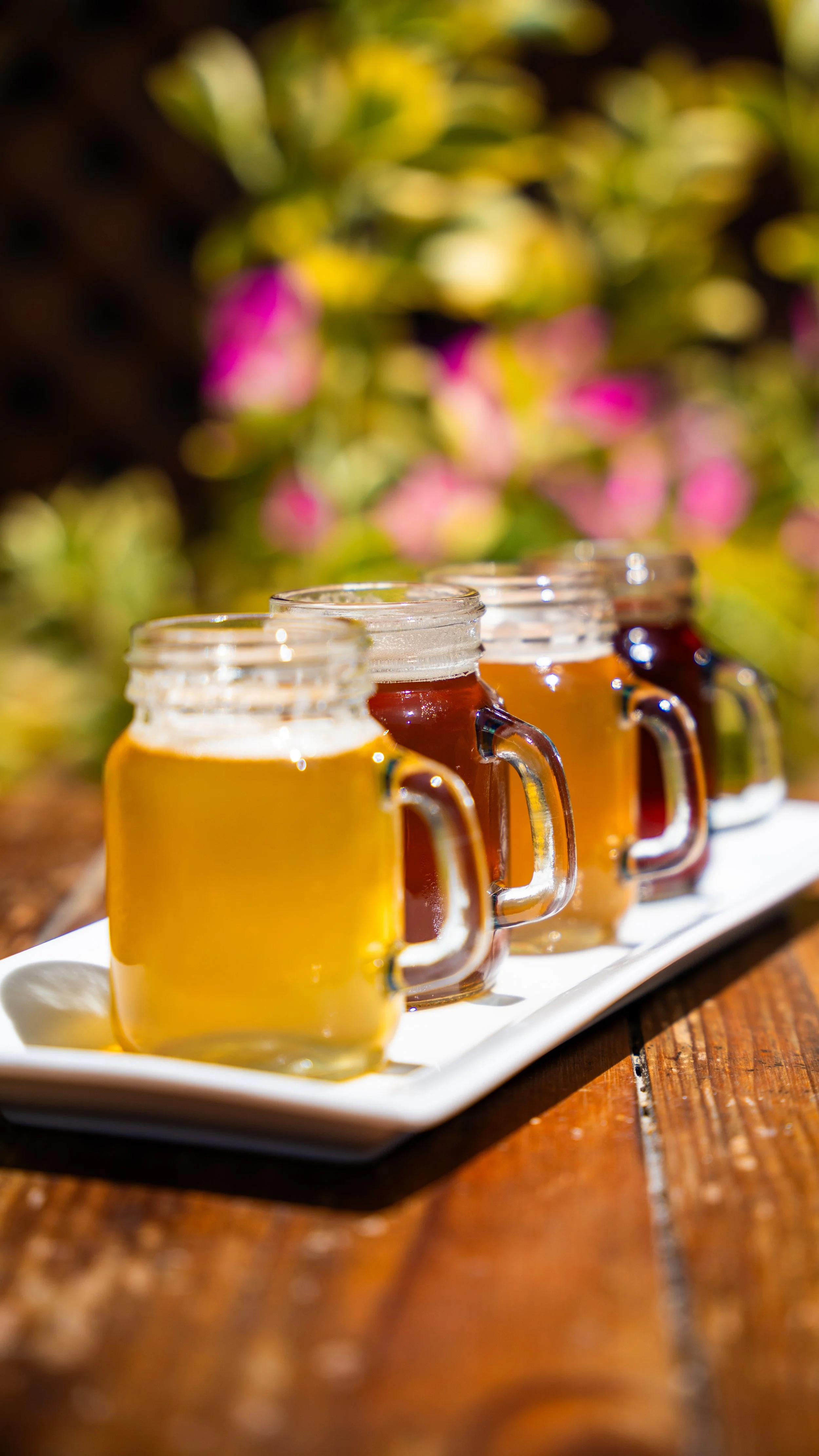 Four small glass mugs filled with different types of beer on a white rectangular plate outdoors, with a blurred background of pink flowers and green foliage.