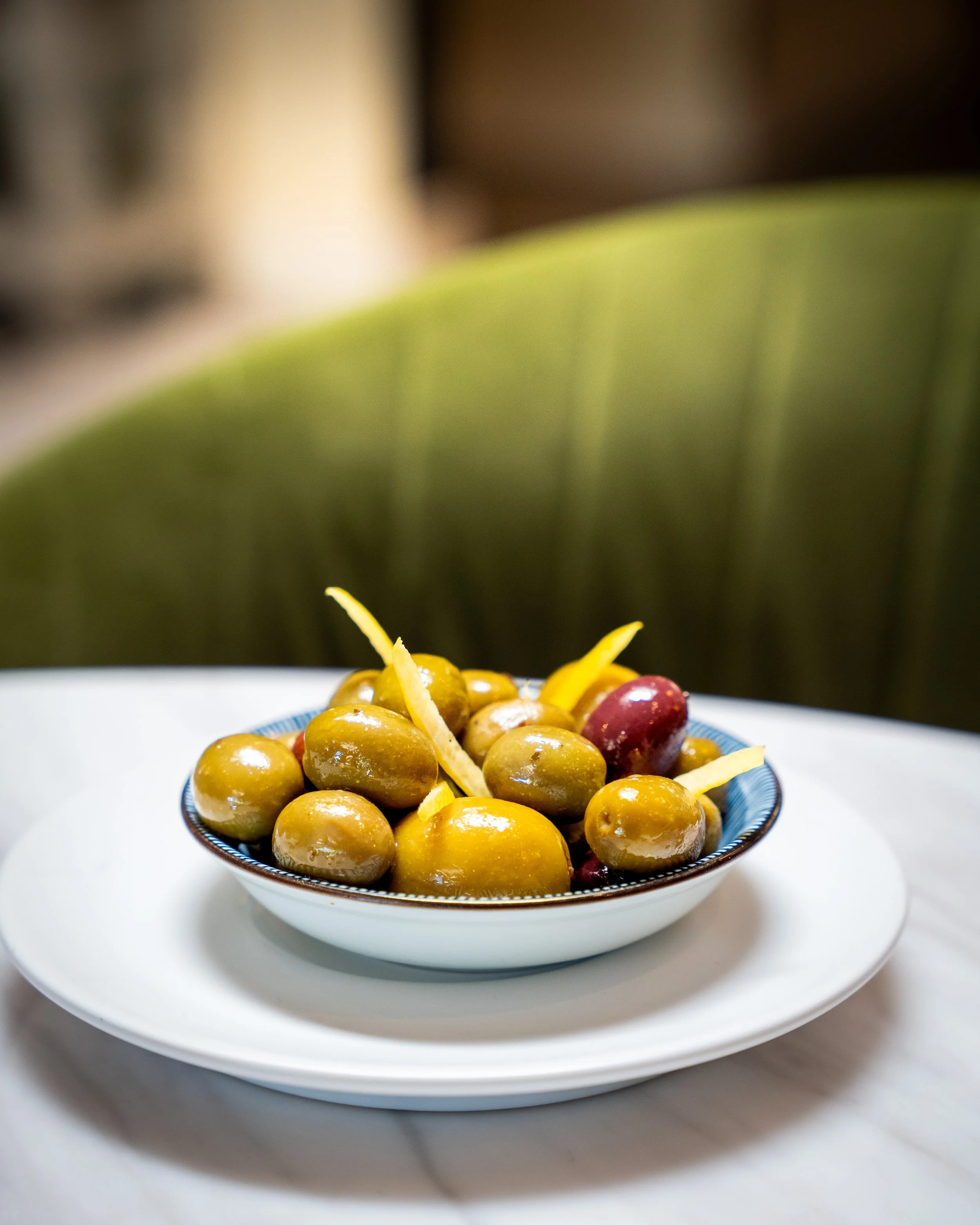A bowl of green olives garnished with lemon zest on a white plate