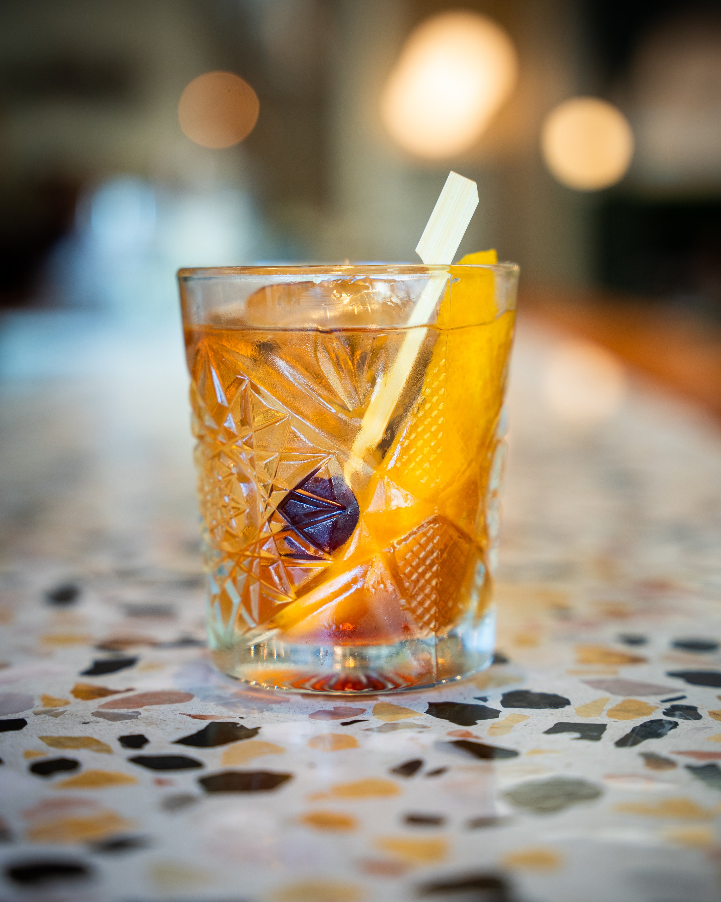 A glass of whiskey with ice, garnished with a lemon wedge and a straw, on a colorful speckled table with blurred warm lights in the background.