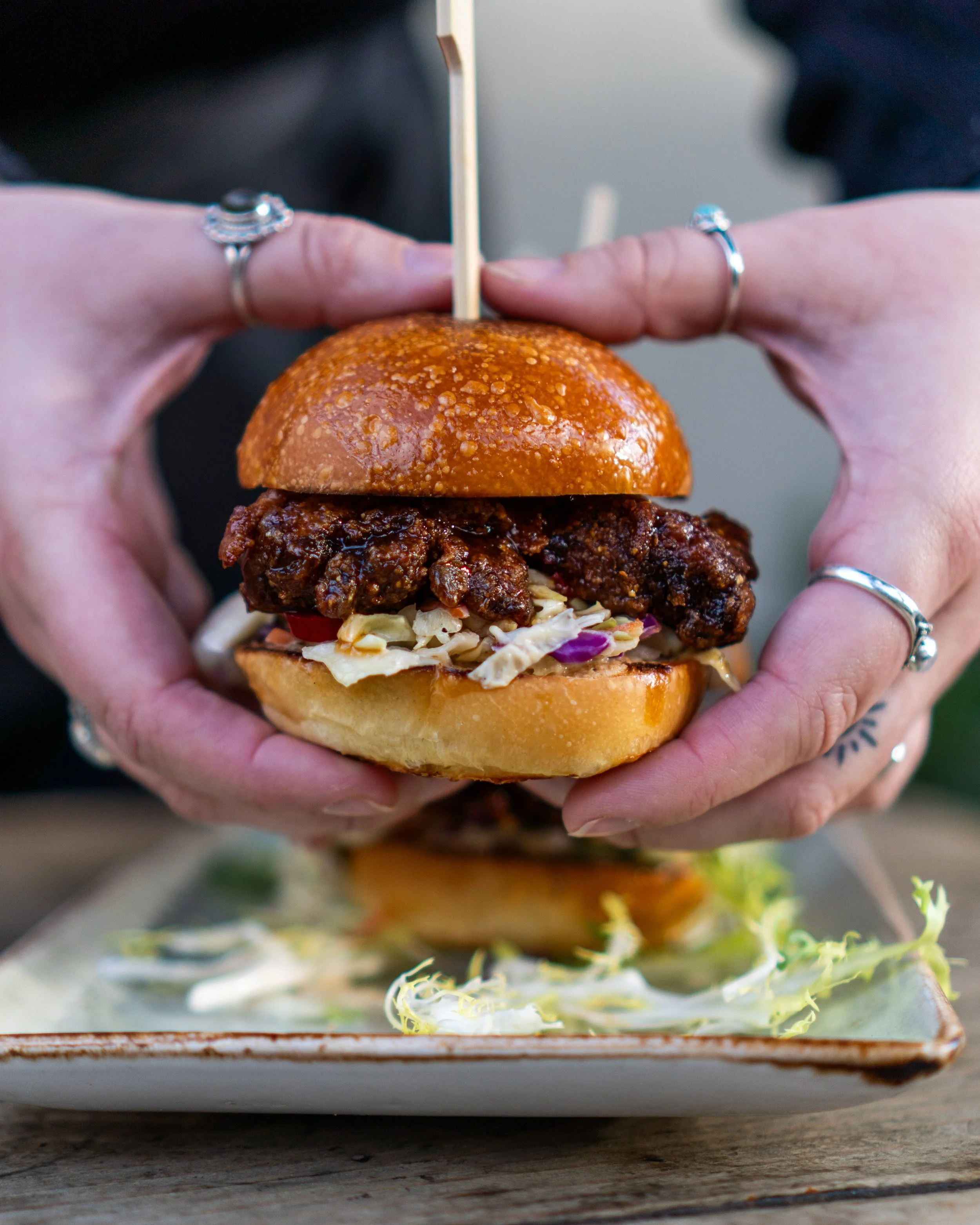 Person holding a fried chicken sandwich with coleslaw, on a bun with a toothpick, on a rectangular plate.