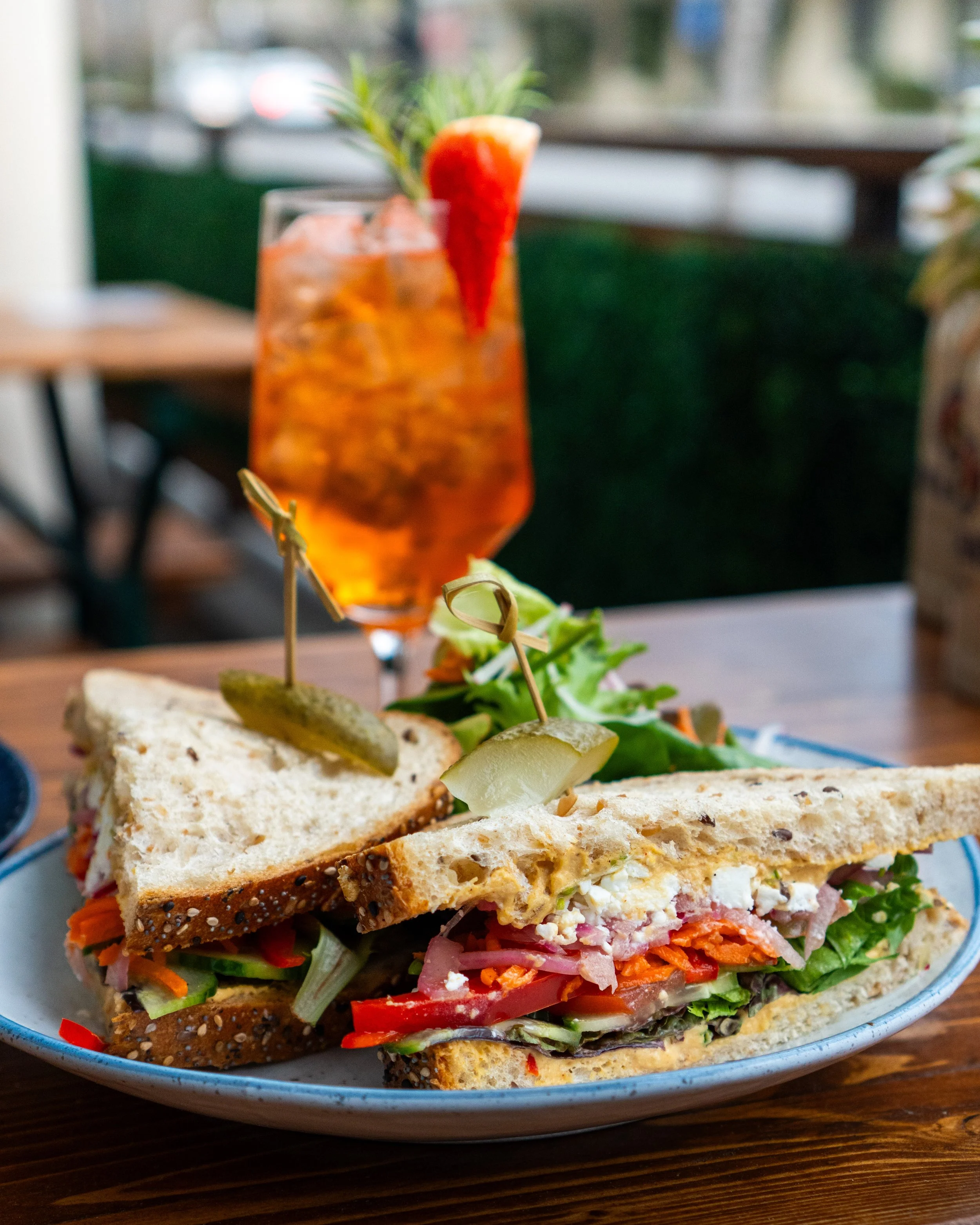 A plate with a sandwich made of multigrain bread, filled with vegetables like lettuce, tomato, carrots, and cheese, garnished with pickle slices and toothpicks, and a glass of iced tea with a strawberry and rosemary garnish on a wooden table