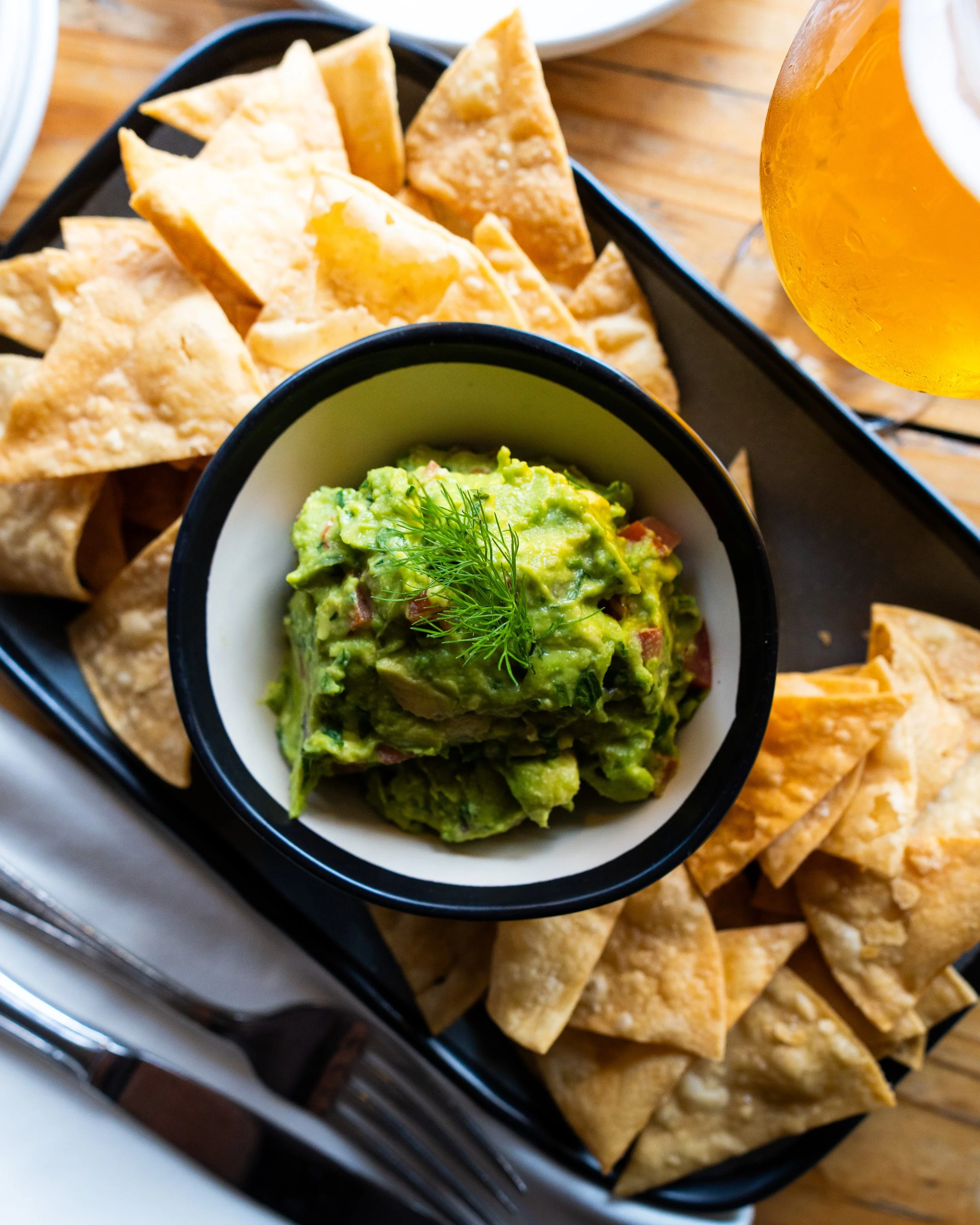 A platter with tortilla chips, a bowl of guacamole topped with dill, and a glass of beer.