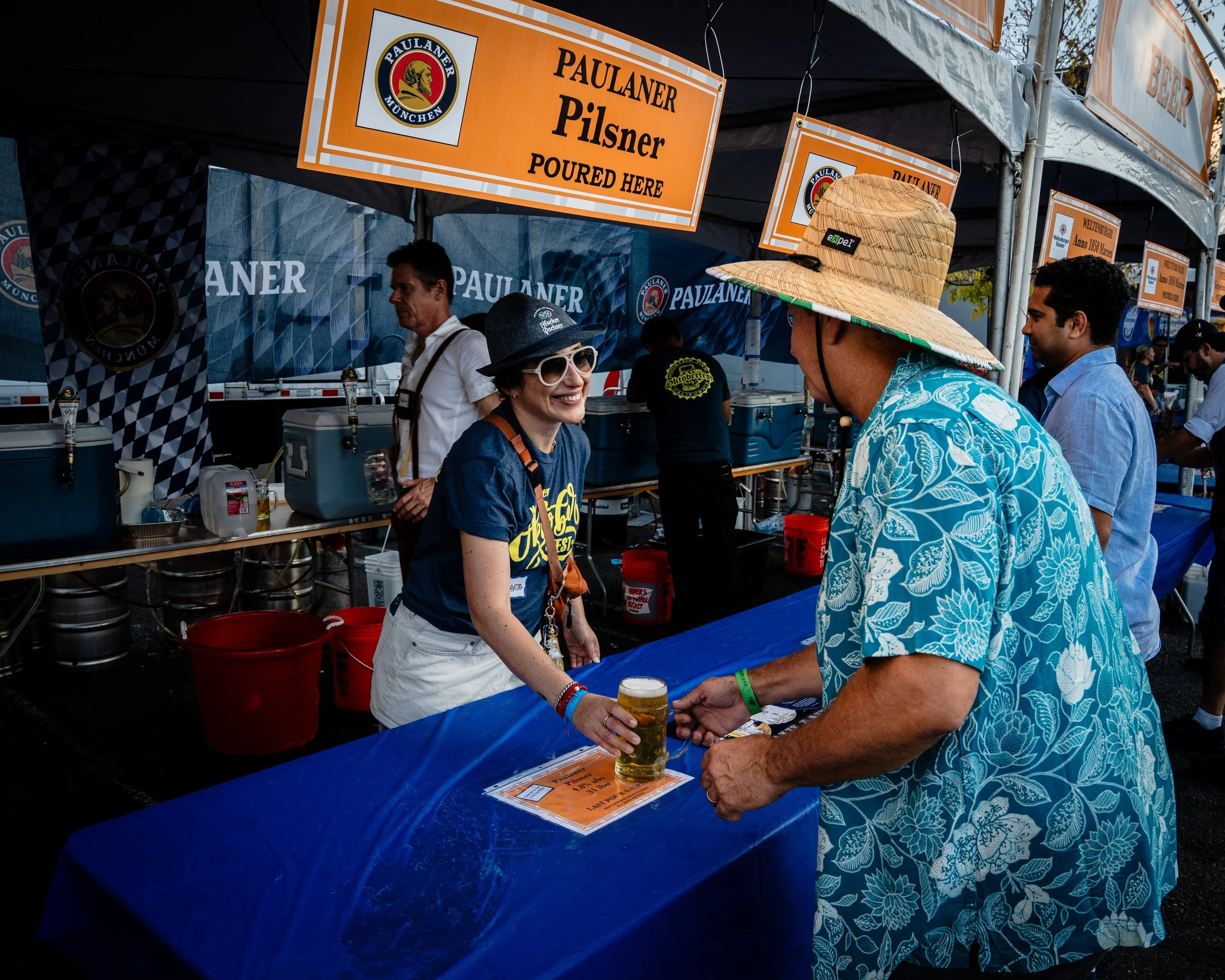 A bartender serving a beer to a customer at an outdoor booth with signs for Paulaner Pilsner, with people in casual summer clothing, including a woman in sunglasses and a man in a Hawaiian shirt and large sunhat.