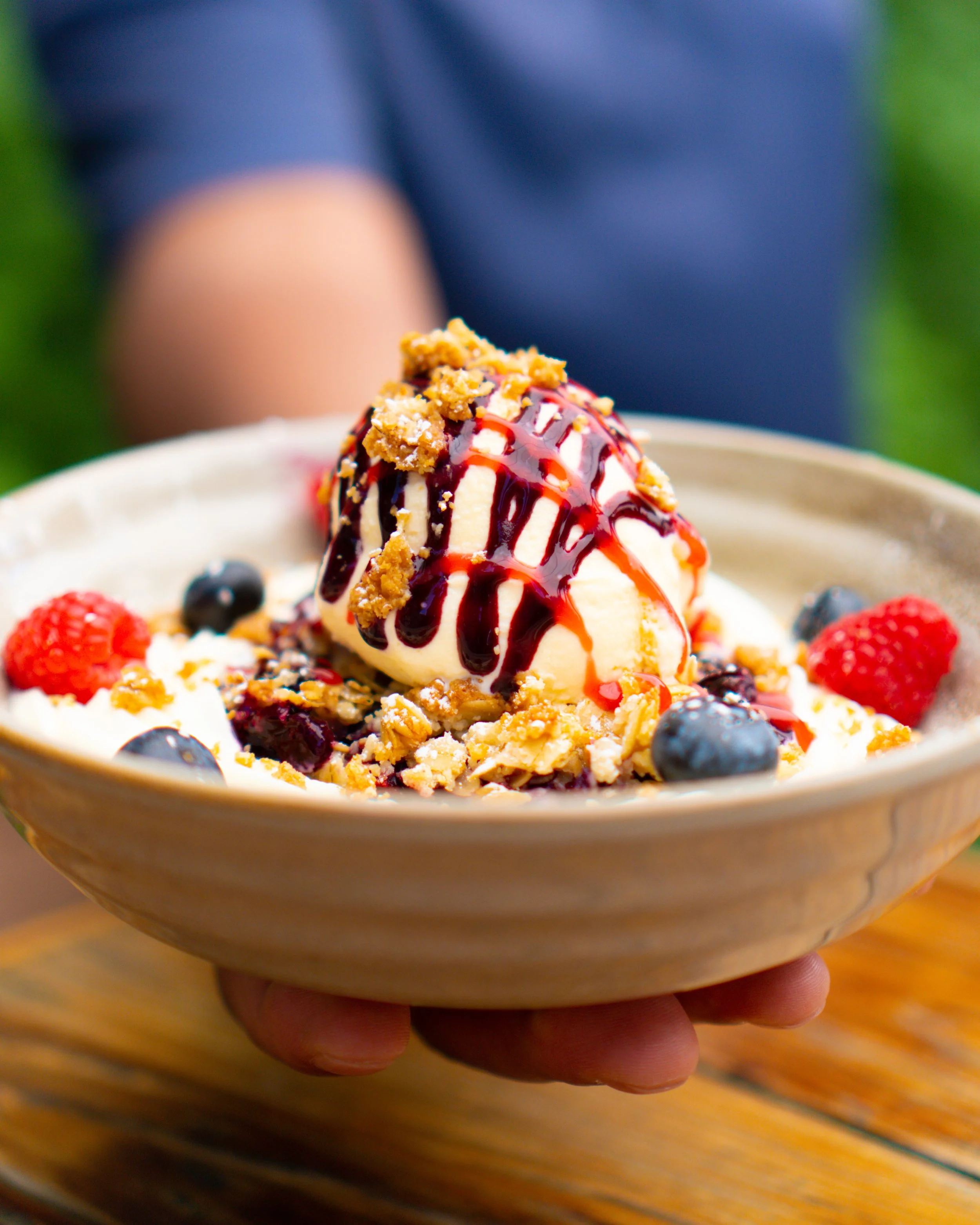 A bowl of vanilla ice cream topped with berry sauce, fresh raspberries and blueberries, and crumbled cookies, held by a person outdoors.