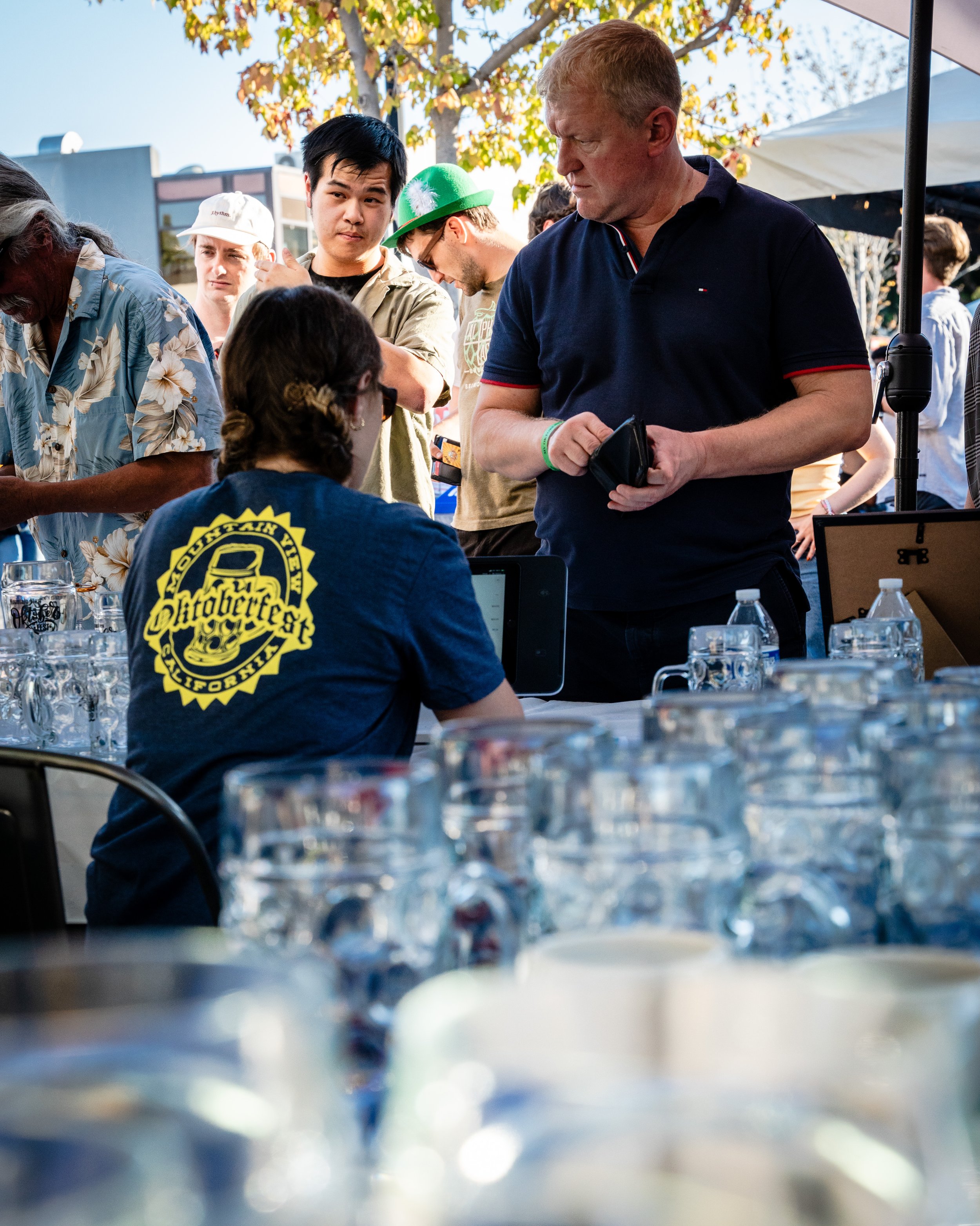 A group of people gathered around a table at an outdoor event. Some are standing, some are sitting. Water glasses are on the table, and a woman in the foreground has a shirt with the word "Oldbikefest" on the back. The scene appears lively with trees