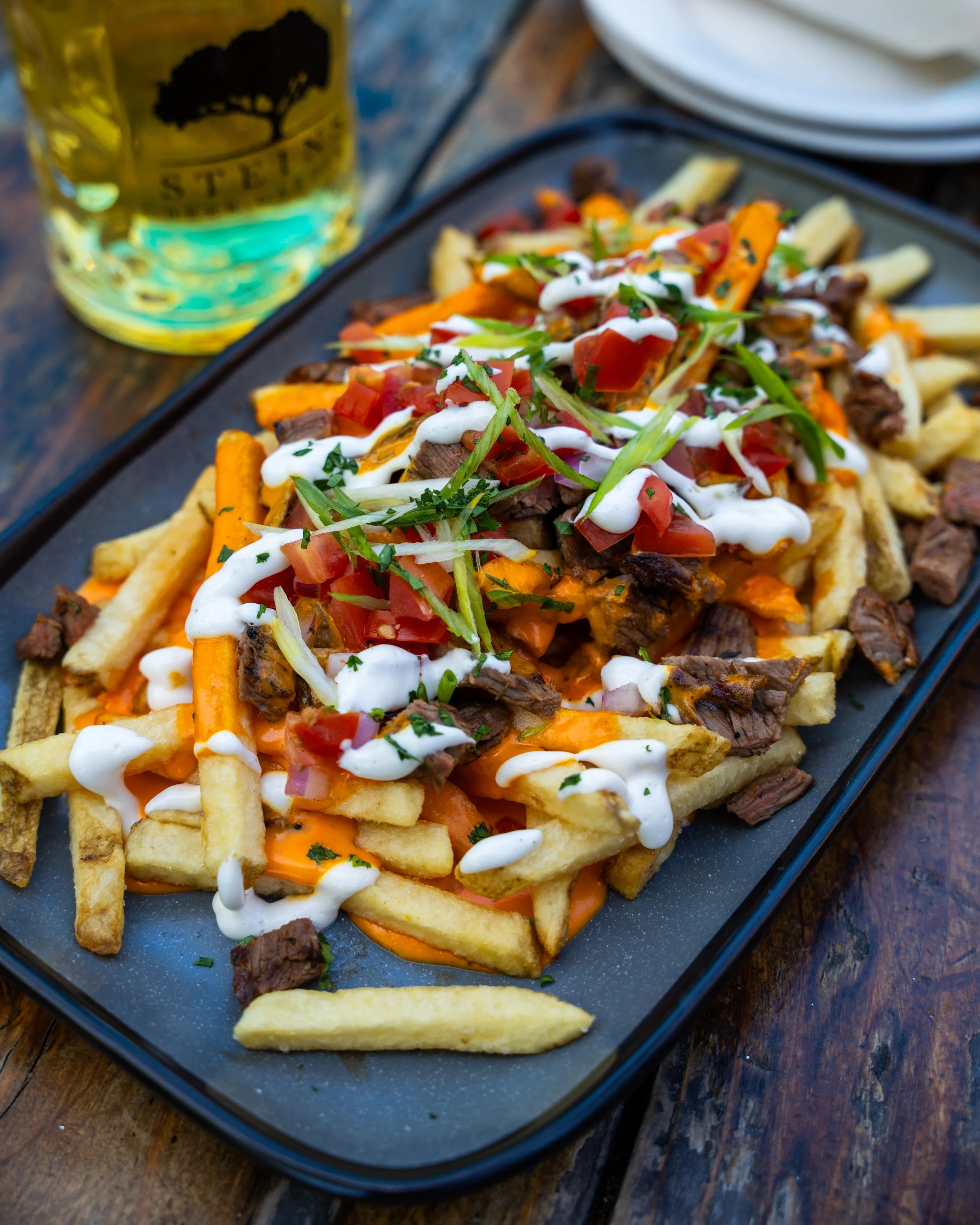A plate of loaded French fries topped with sliced steak, diced tomatoes, shredded cheese, chopped onions, green herbs, and drizzled with white sauce, with a glass of beer nearby at restaurant in DMV area.