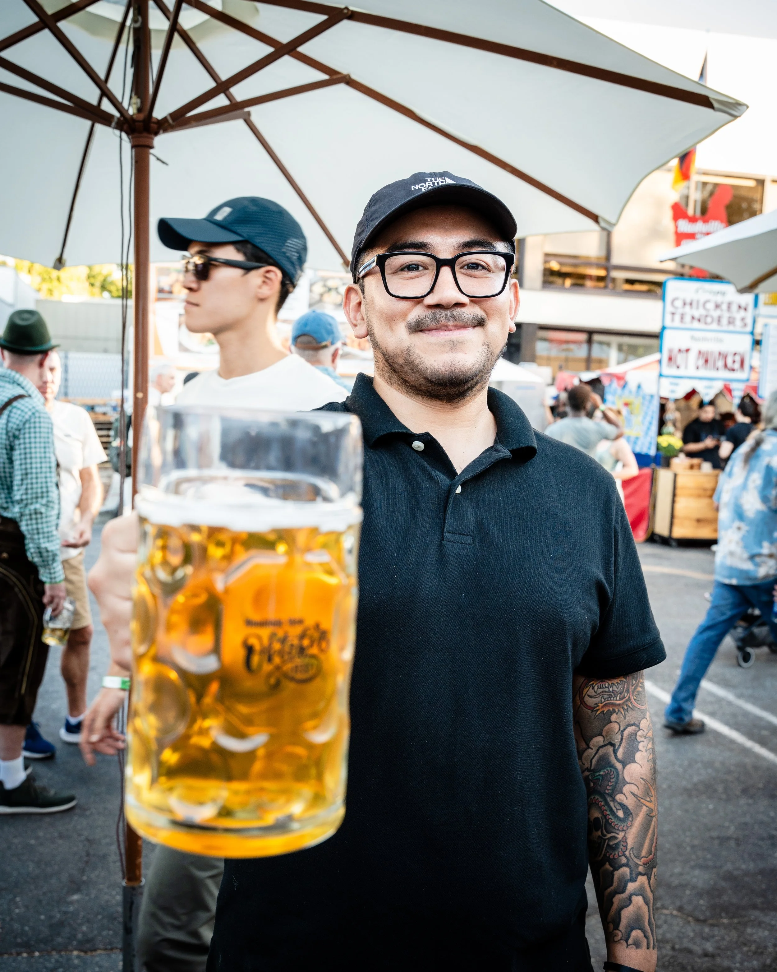 A man smiling and holding a glass of beer at an outdoor festival or fair. He is wearing glasses, a black polo shirt, a cap, and has a tattooed arm. There are tents, signs, and other people in the background. At a brewery in Maryland. 