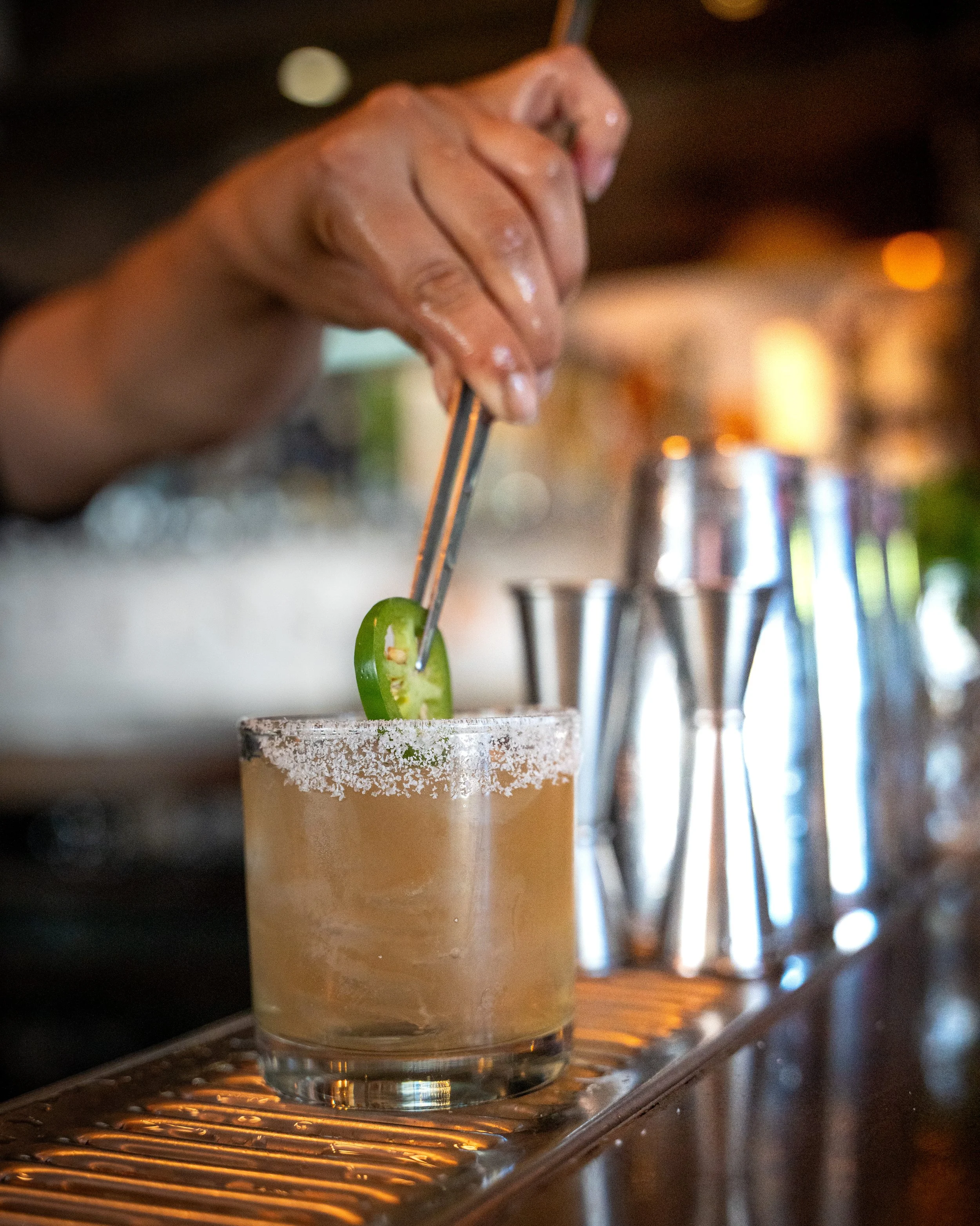 A bartender garnishing a cocktail with a slice of jalapeño pepper, using tongs, at a bar in Washington DC.