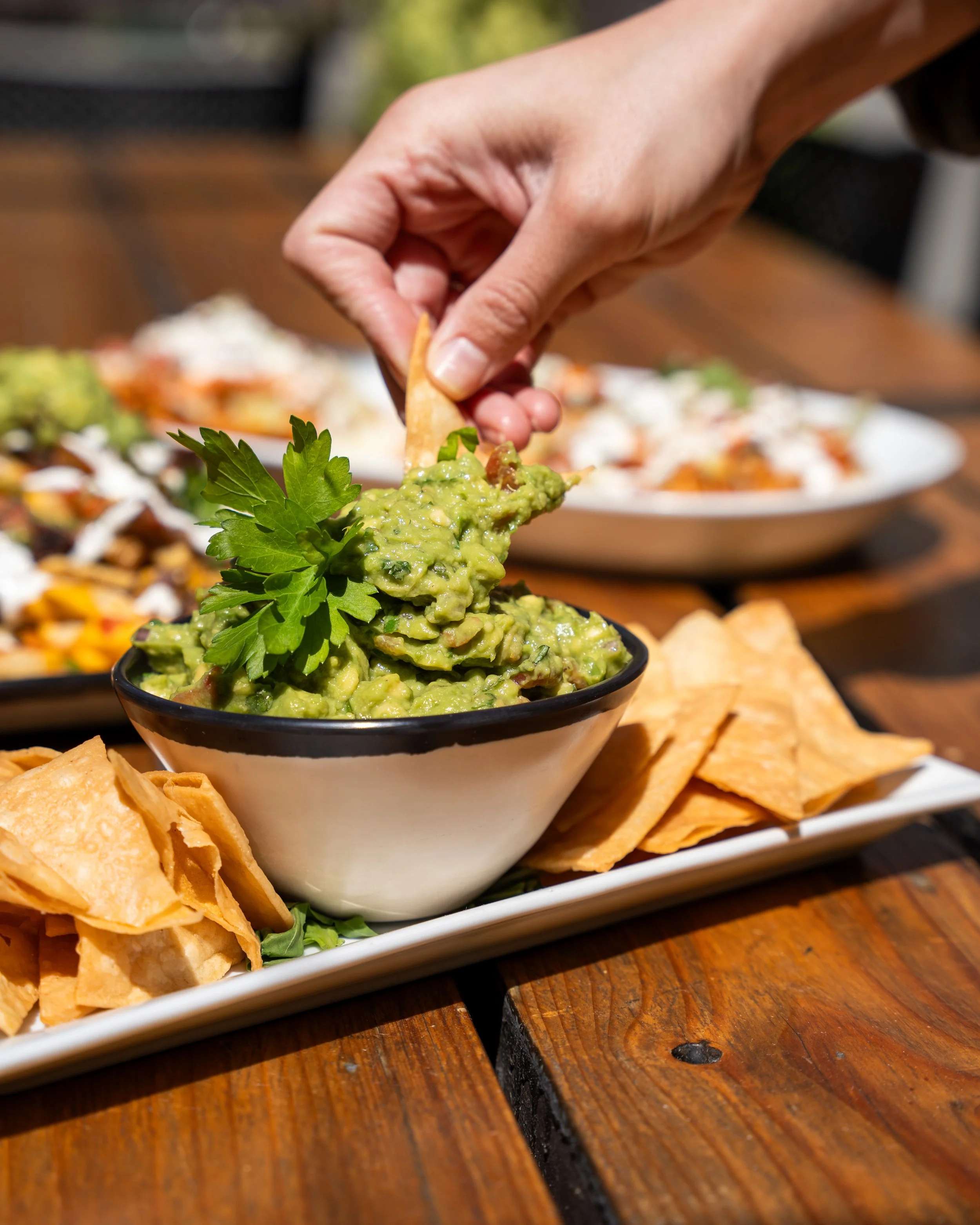 A hand dipping a chip into a bowl of guacamole garnished with a sprig of parsley, with tortilla chips on the side and blurred plates of food in the background on a wooden table.