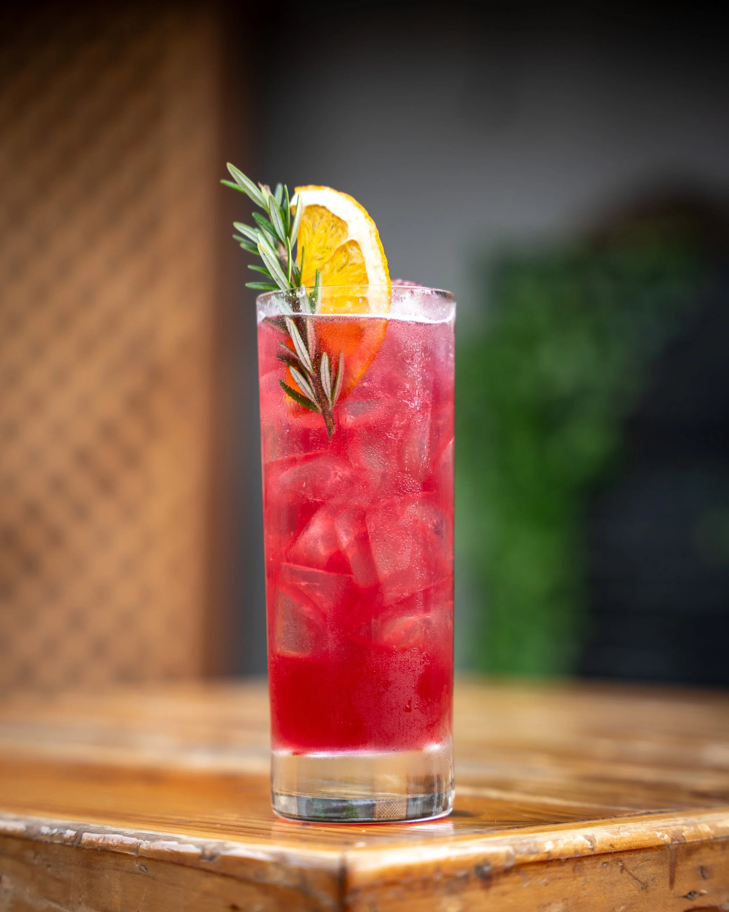 A tall glass of red cocktail garnished with a slice of orange and a sprig of rosemary, with ice cubes inside.