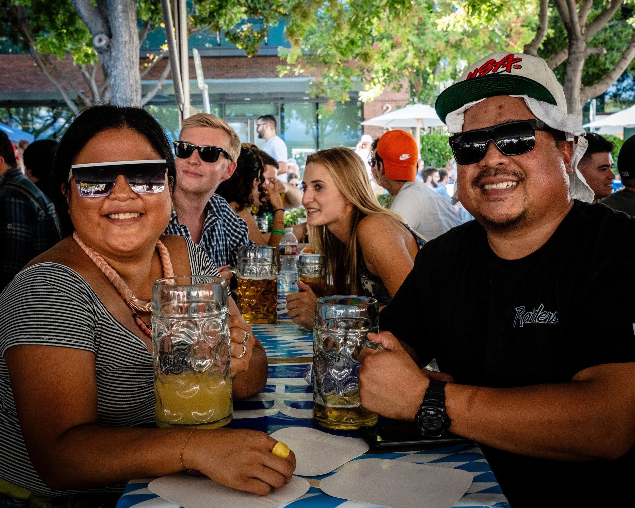 Group of people enjoying drinks at an outdoor gathering, smiling, wearing sunglasses, with beer mugs on a table with a blue and white geometric tablecloth, trees, and a building in the background.