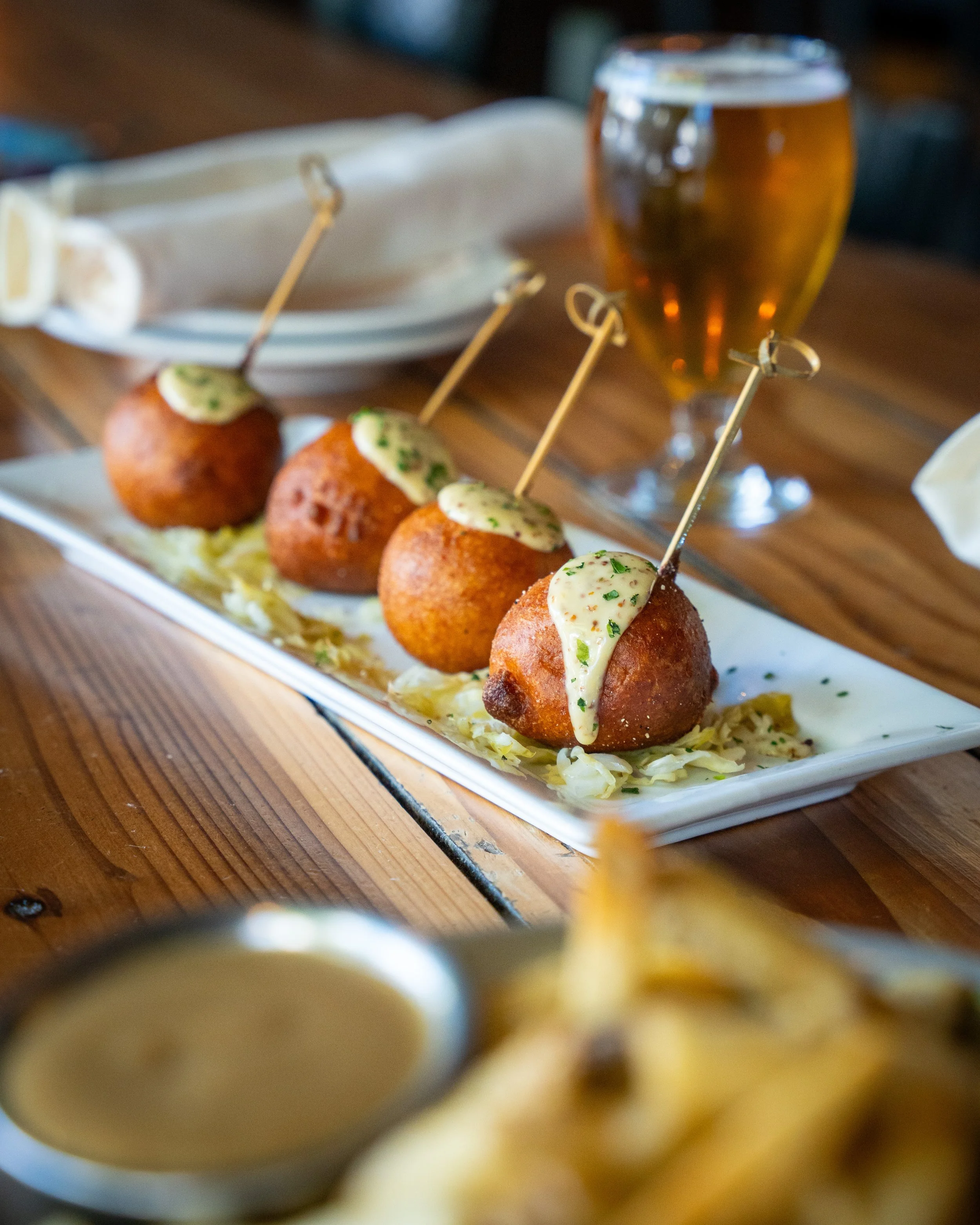 Four fried appetizers with a creamy herb sauce on a rectangular white plate, set on a wooden table with a glass of beer in the background in Maryland restaurant.